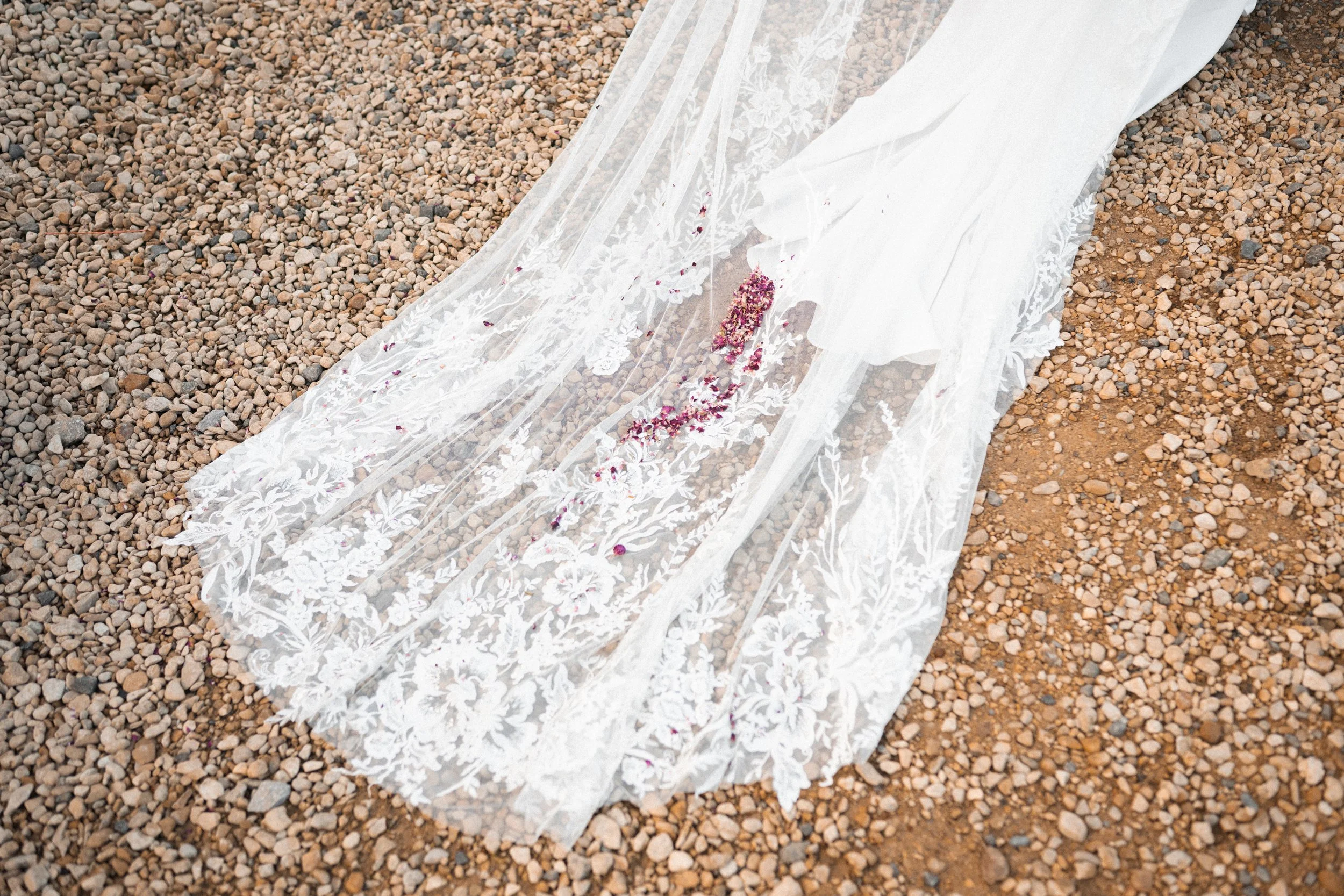 Wedding dress with lace and embroidery laid on gravel ground with scattered pink flower petals.