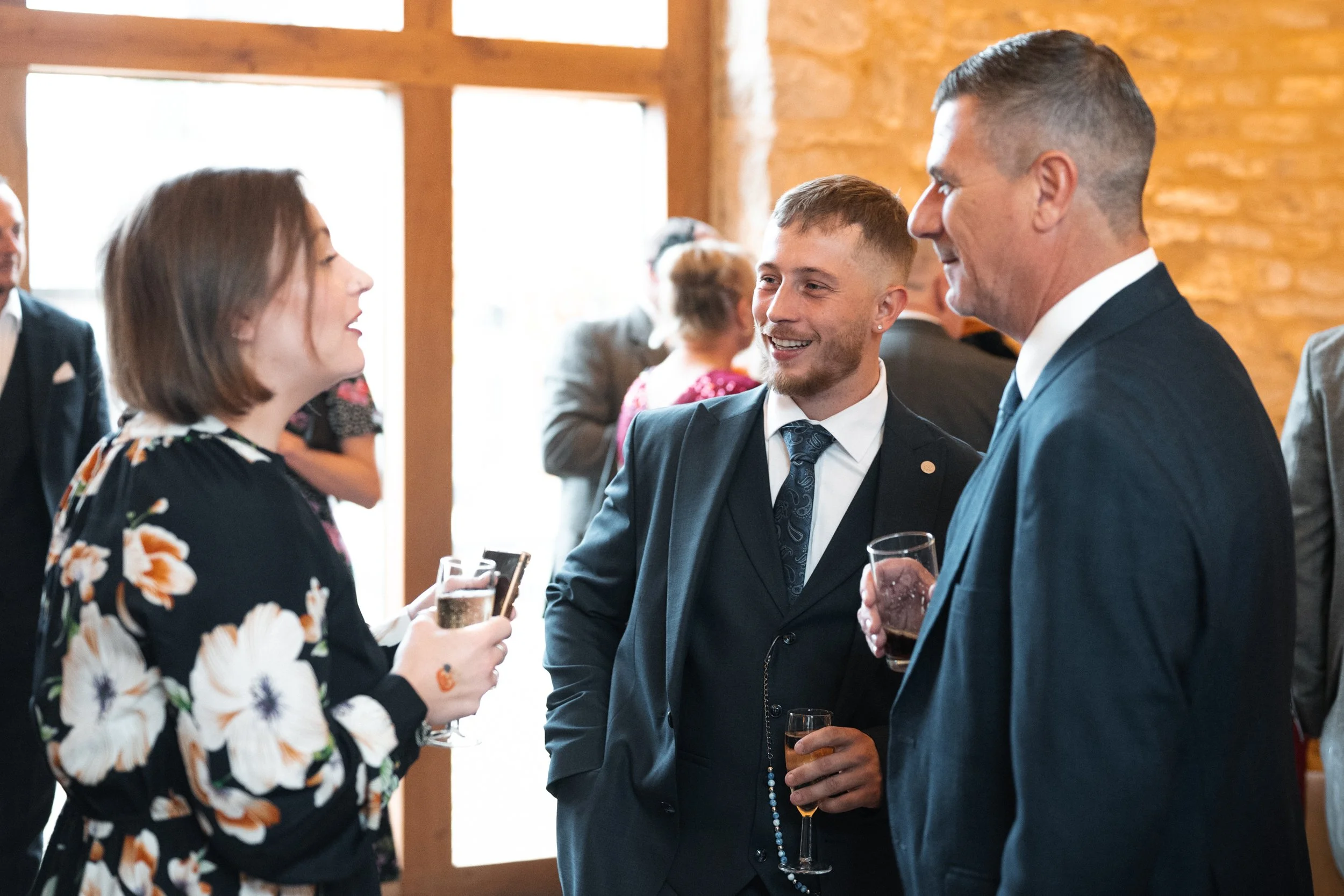 Three people in formal attire, two men and one woman, engaged in conversation at a social event, holding drinks in a warmly lit room with wooden walls.