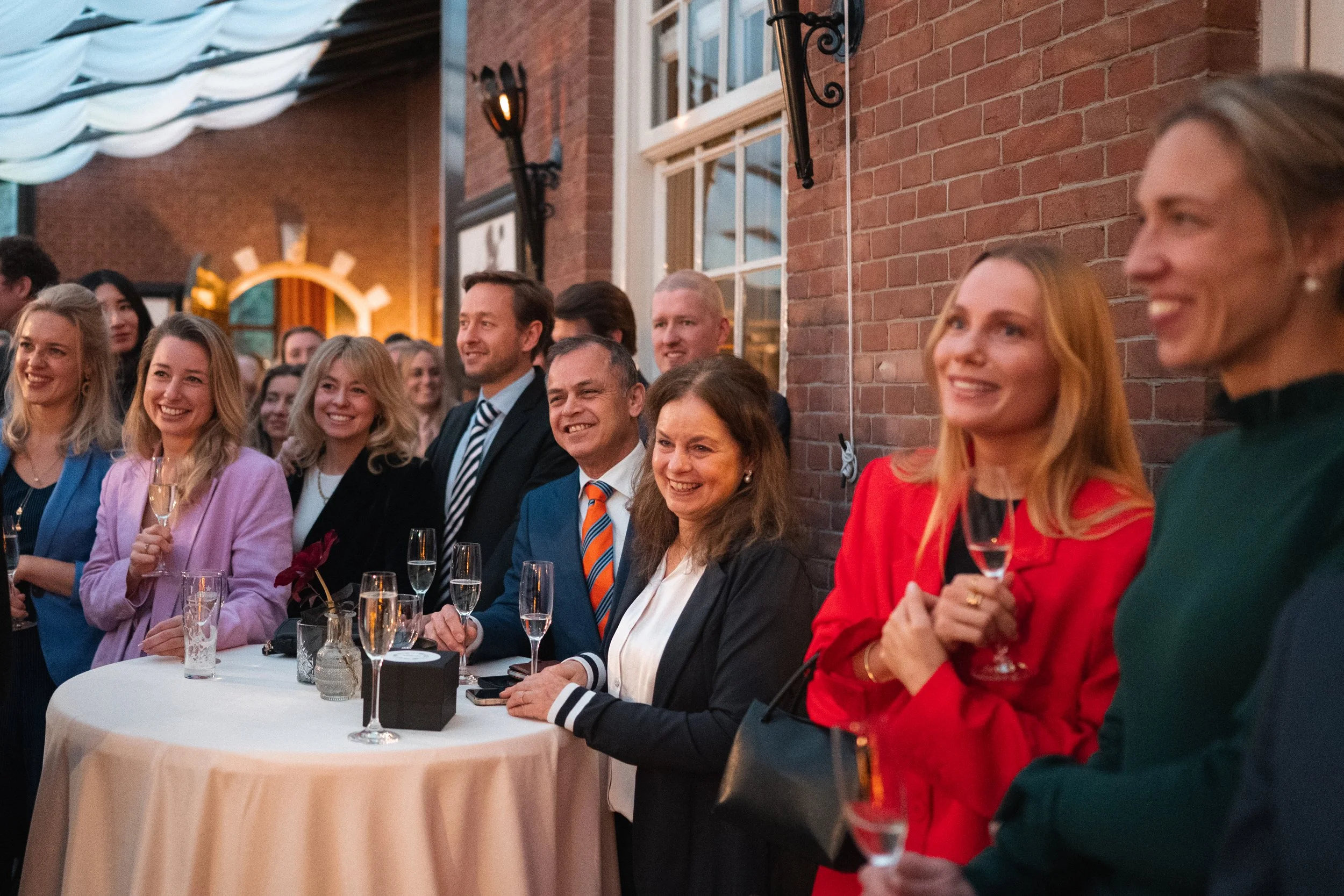 People smiling and holding glasses of champagne at a social gathering or celebration inside a venue with exposed brick walls.