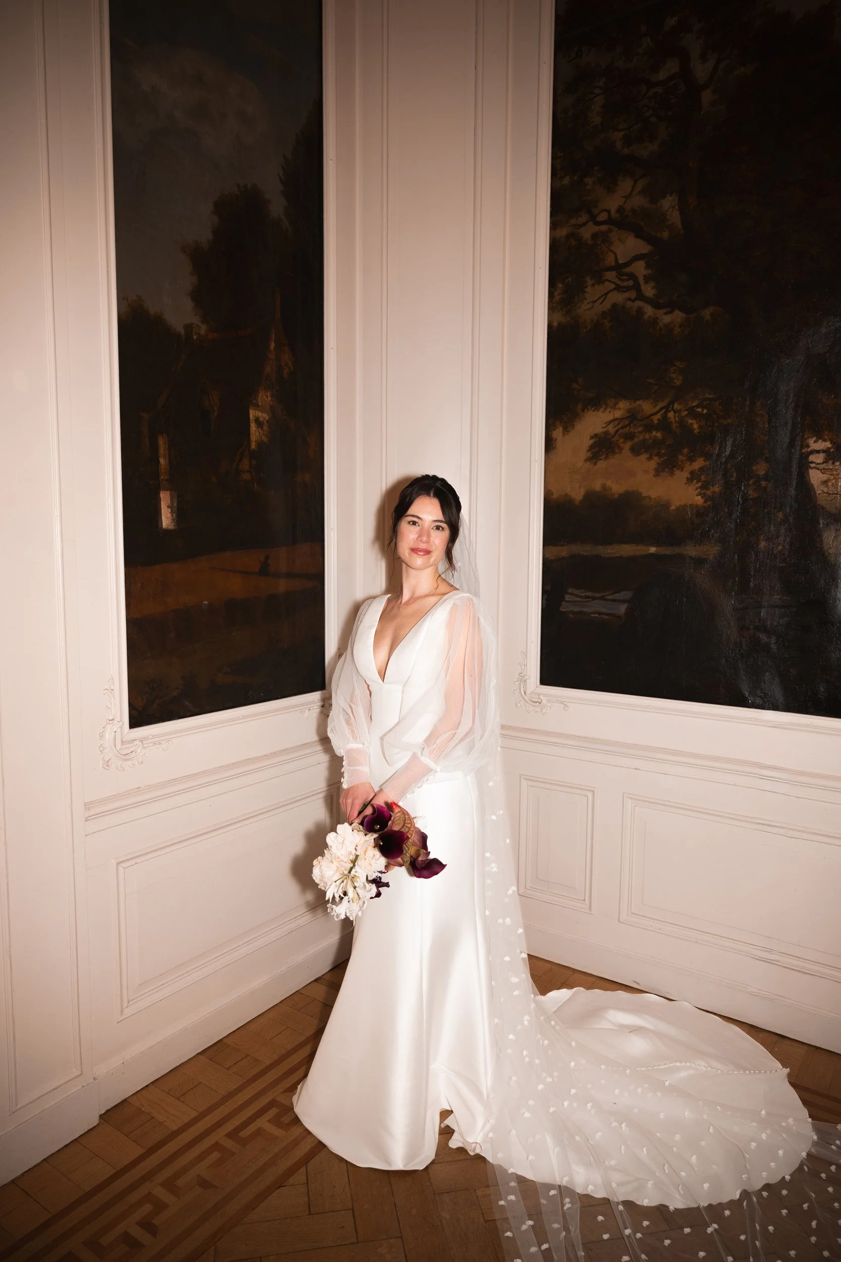 A woman in a white wedding dress holding a bouquet of flowers stands inside a room with white walls and large framed mural paintings of trees outside at night.