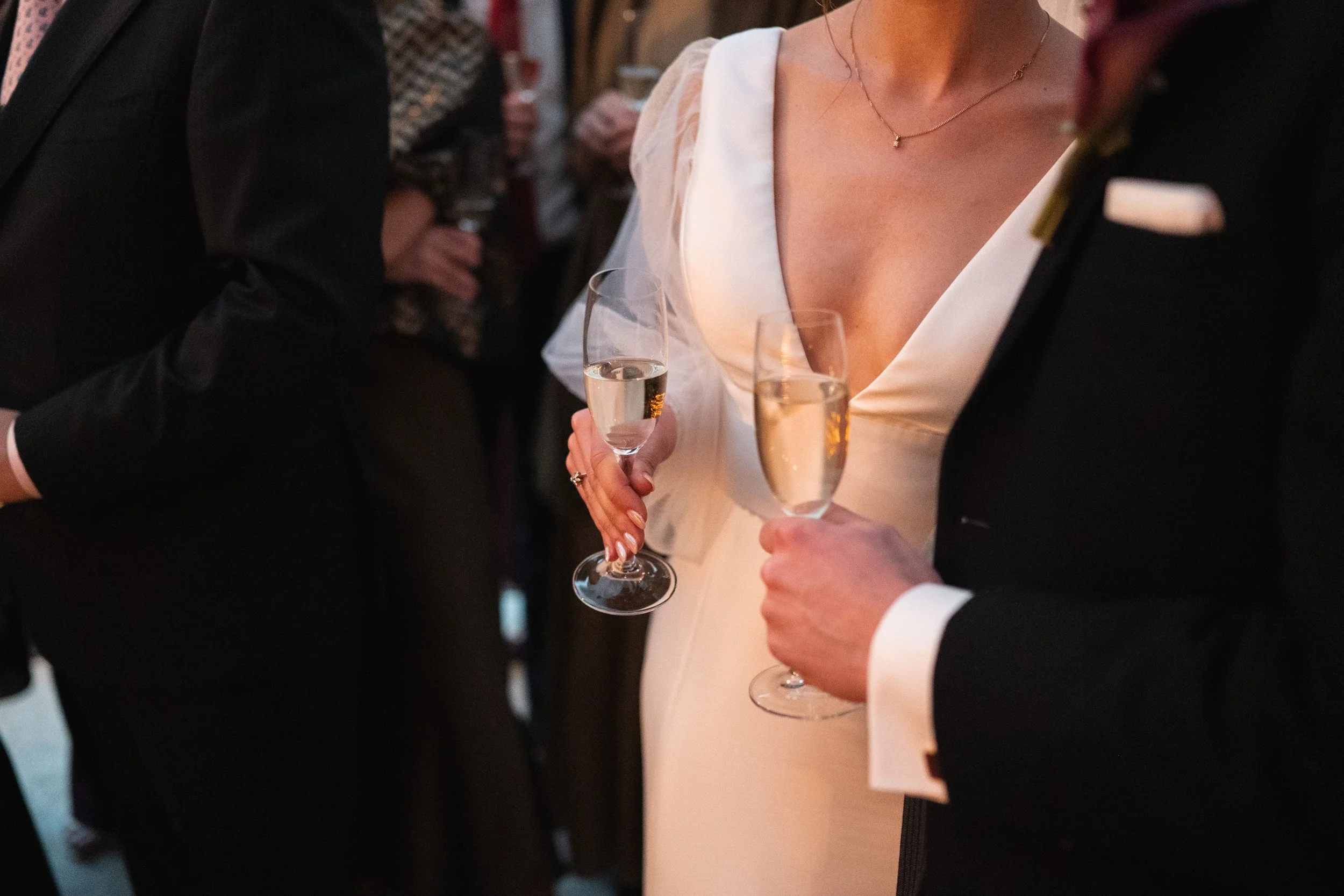 Close-up of a woman in a white dress holding a champagne flute, with a man in a tuxedo also holding a champagne flute, at an indoor celebration or wedding event.