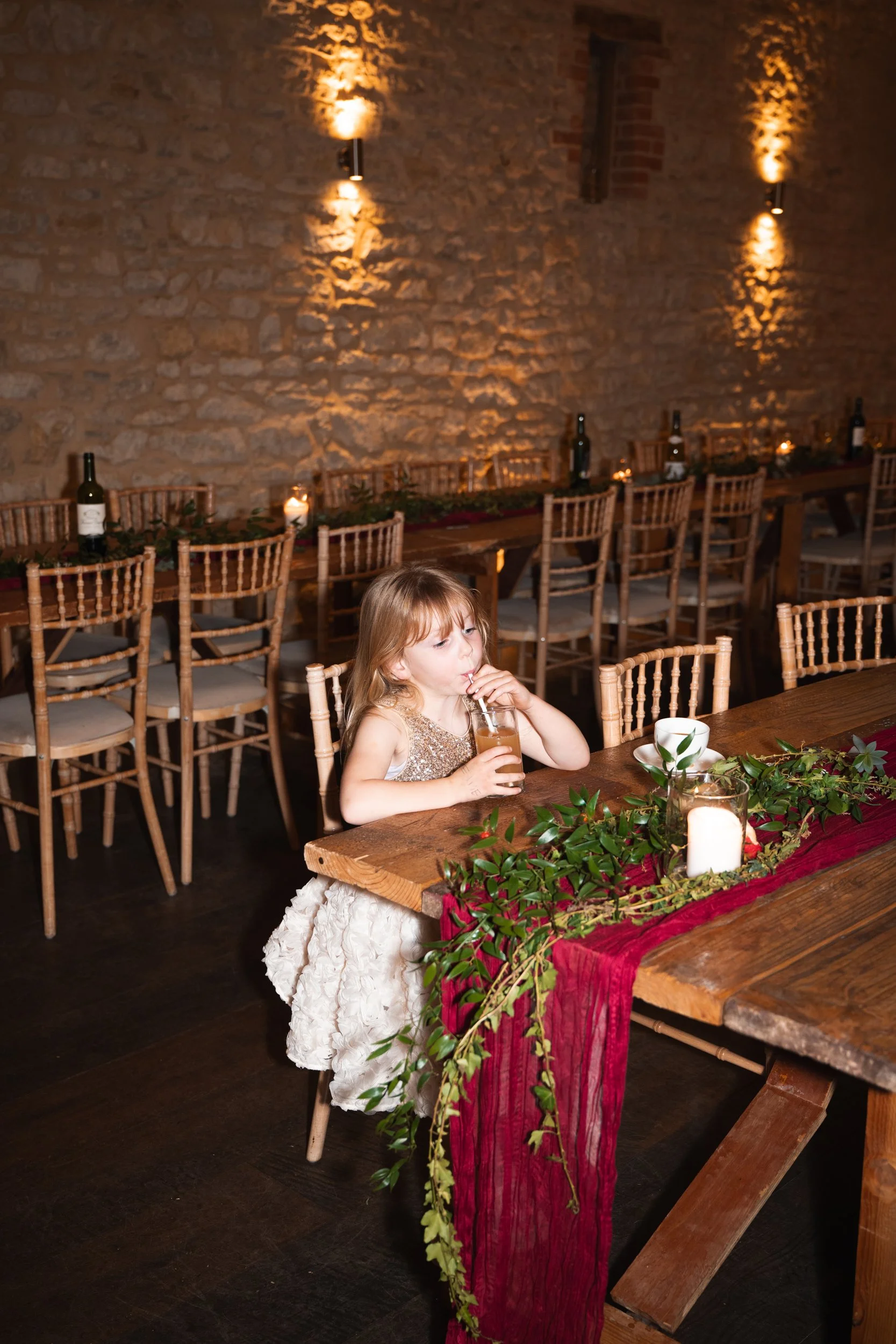 A young girl with long blonde hair sitting at a large wooden table, drinking from a glass with a straw. The table is decorated with greenery, a candle, and a red cloth runner. The setting appears to be a rustic restaurant with exposed brick walls and