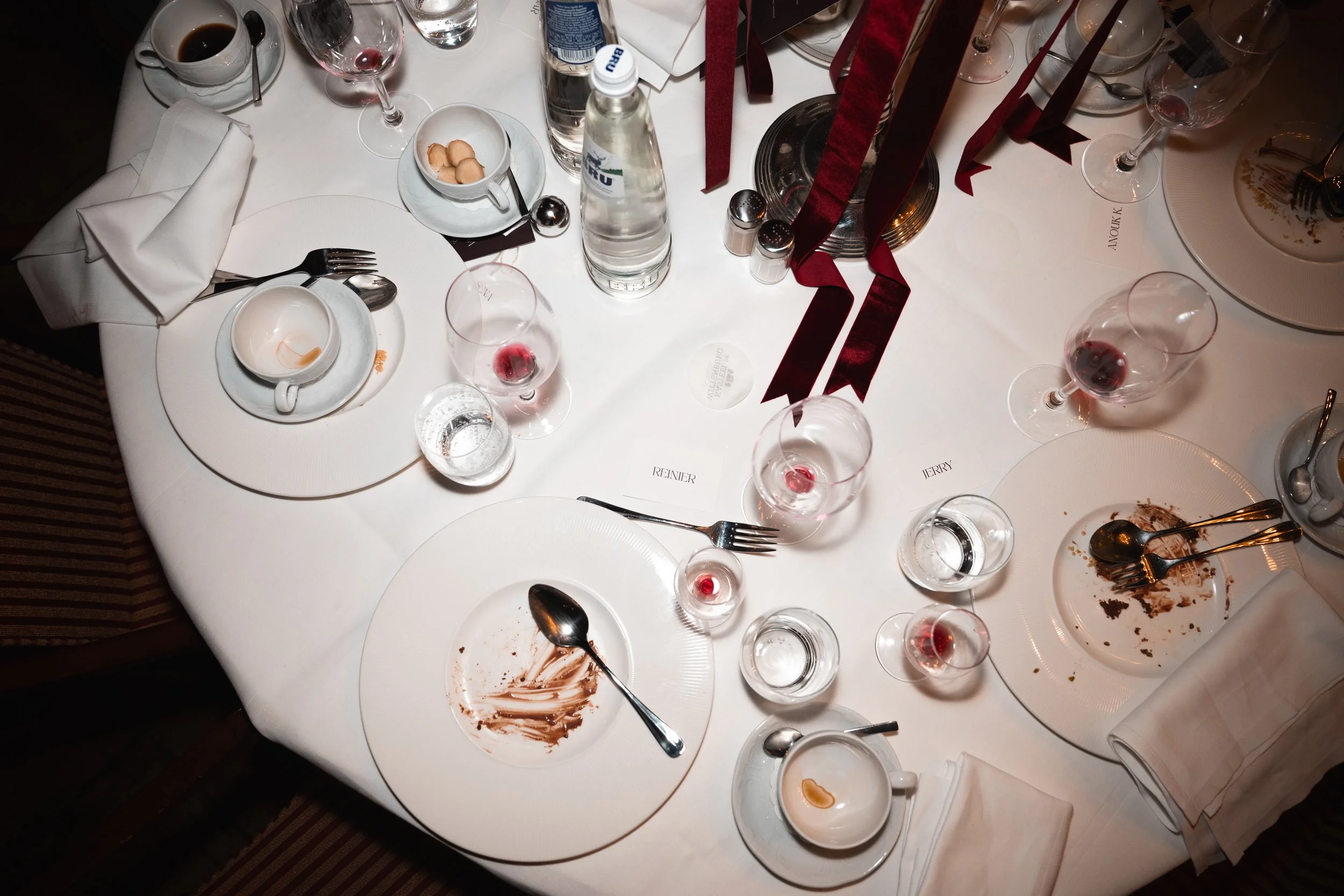 Empty and used dinner plates, glasses with red wine, water bottles, utensils, and leftover food crumbs on a white tablecloth at a formal event.