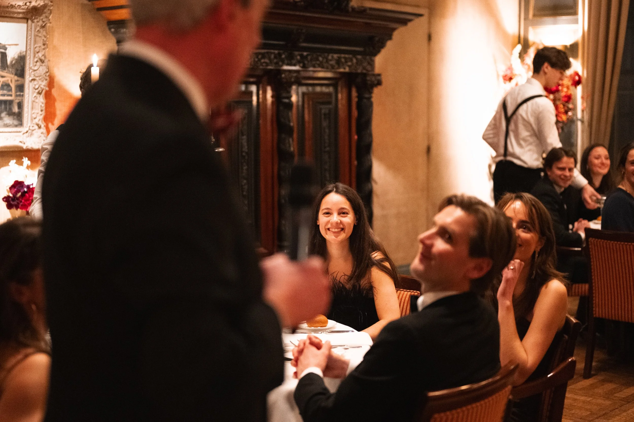 People dressed in formal attire, seated at a dinner table, smiling and engaging in conversation at an elegant restaurant or banquet hall.