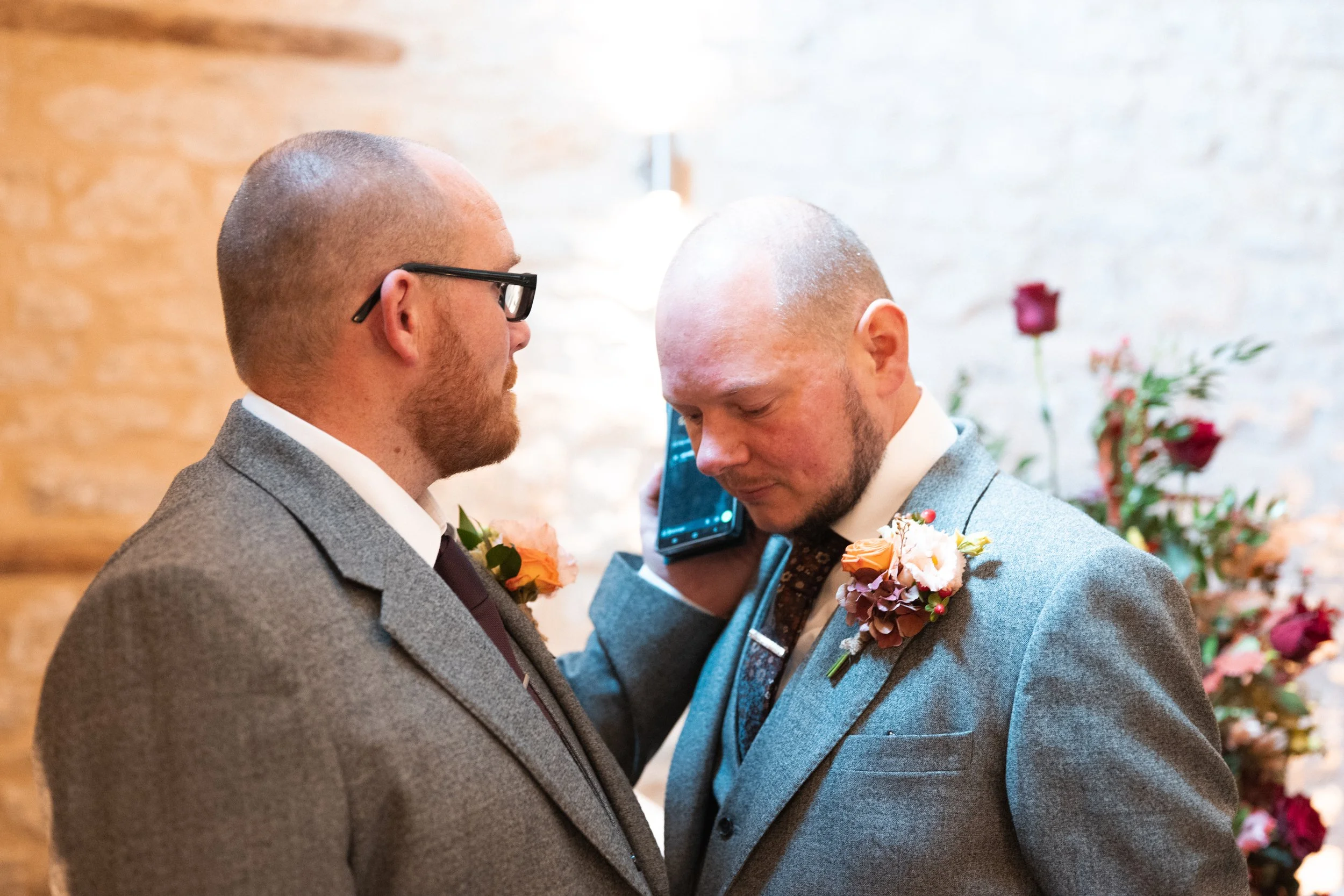 Two men in suits with boutonnieres, one holding a phone to his ear, close together with serious expressions at a wedding or formal event.