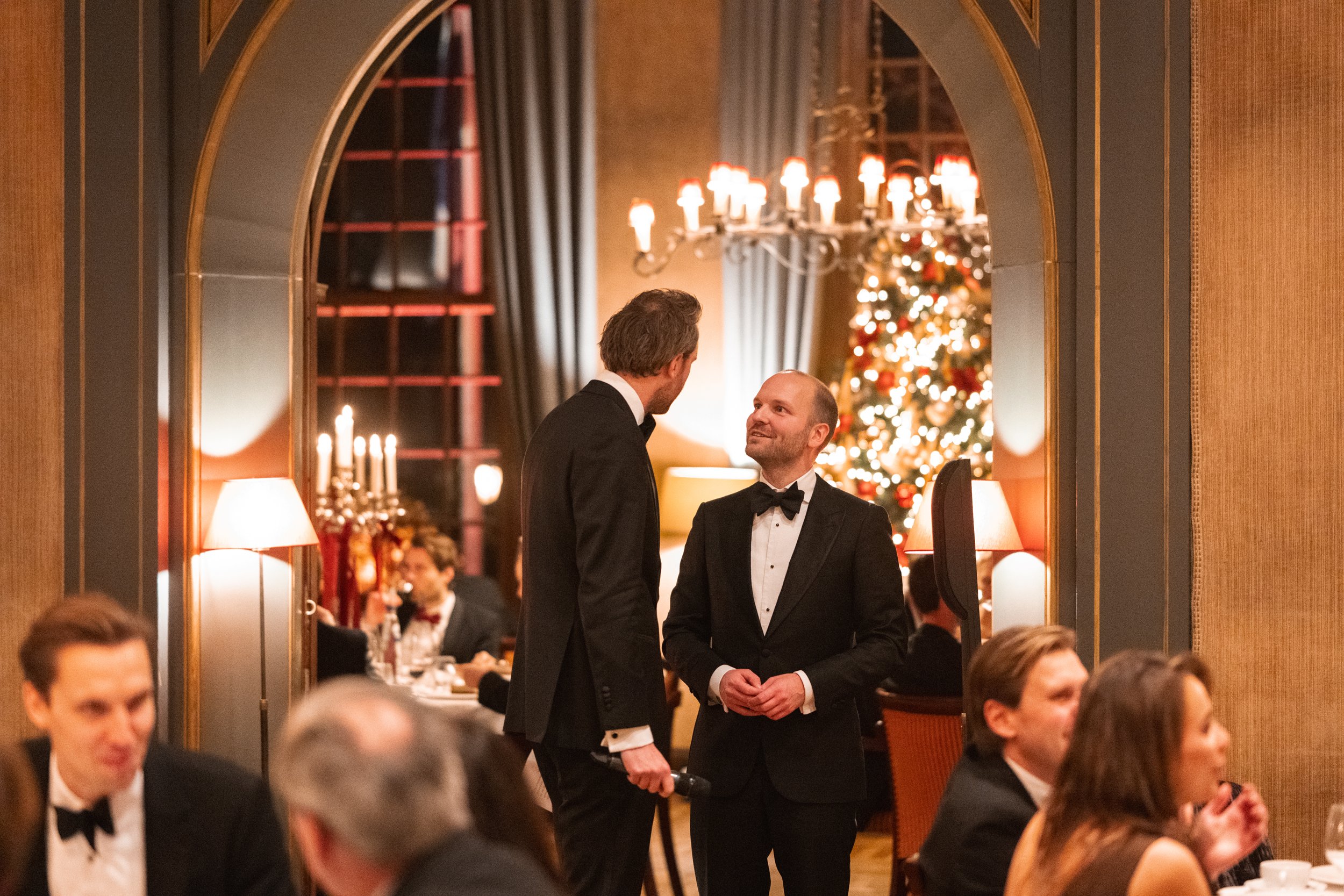 Two men in tuxedos talking at a formal dinner event with a decorated Christmas tree in the background.
