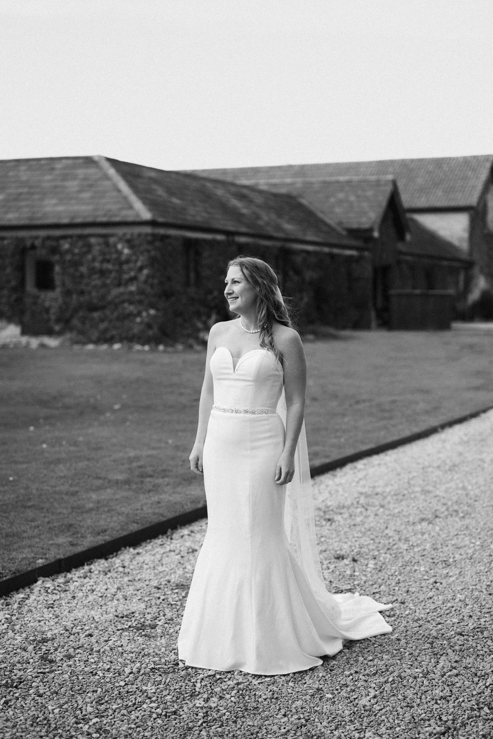 A woman in a white wedding dress standing outdoors on a gravel path, smiling, with buildings in the background.