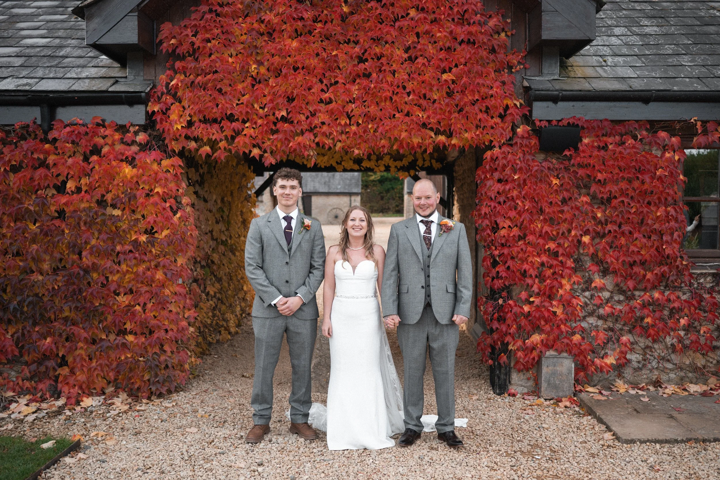 A wedding couple and a man standing hand in hand under an archway covered in red and orange autumn ivy leaves.