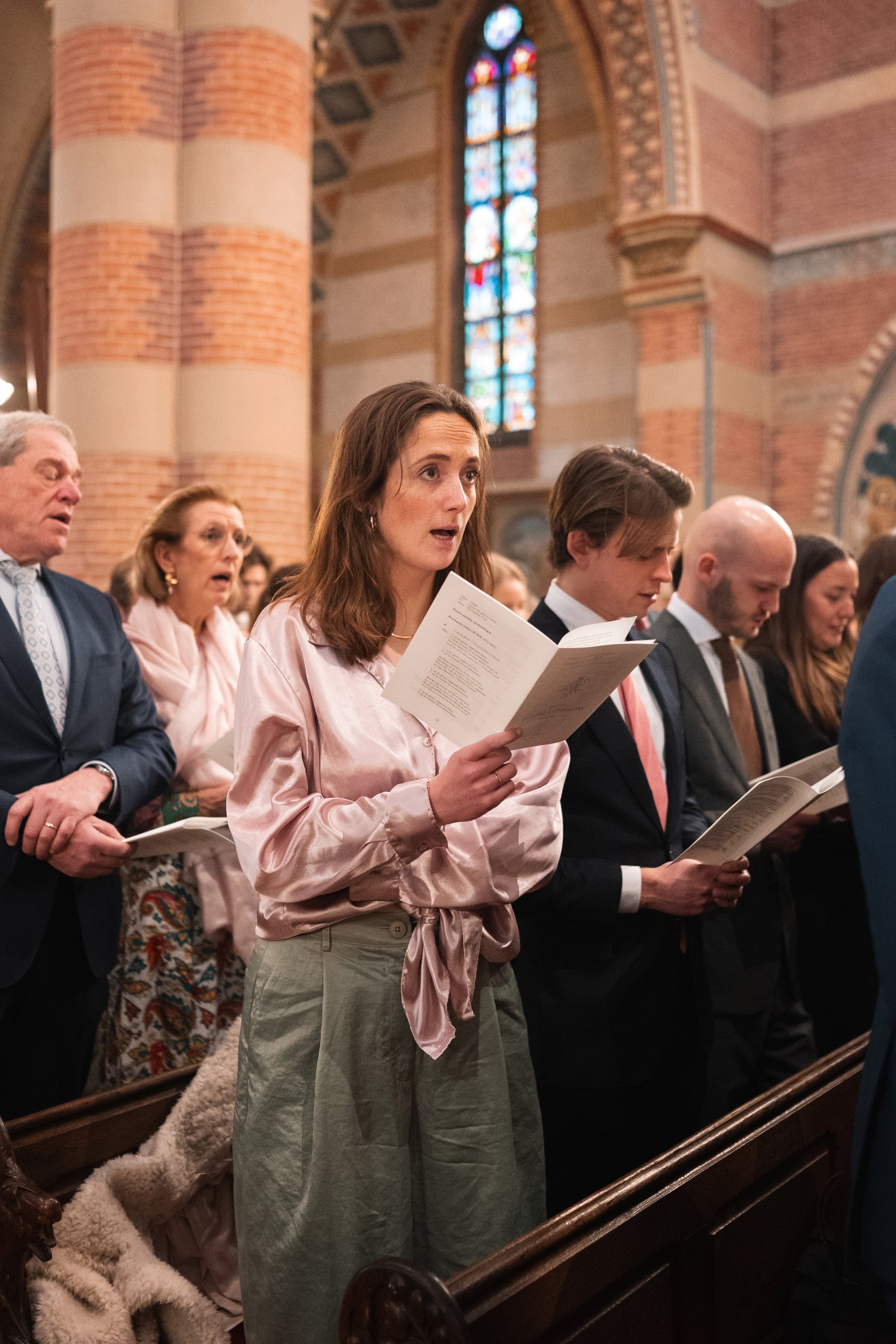 A group of people standing in a church, holding hymnbooks and singing. The woman in the foreground has shoulder-length brown hair, is wearing a pink satin blouse with a bow at the waist, and beige pants. The church has brick walls, stained glass wind