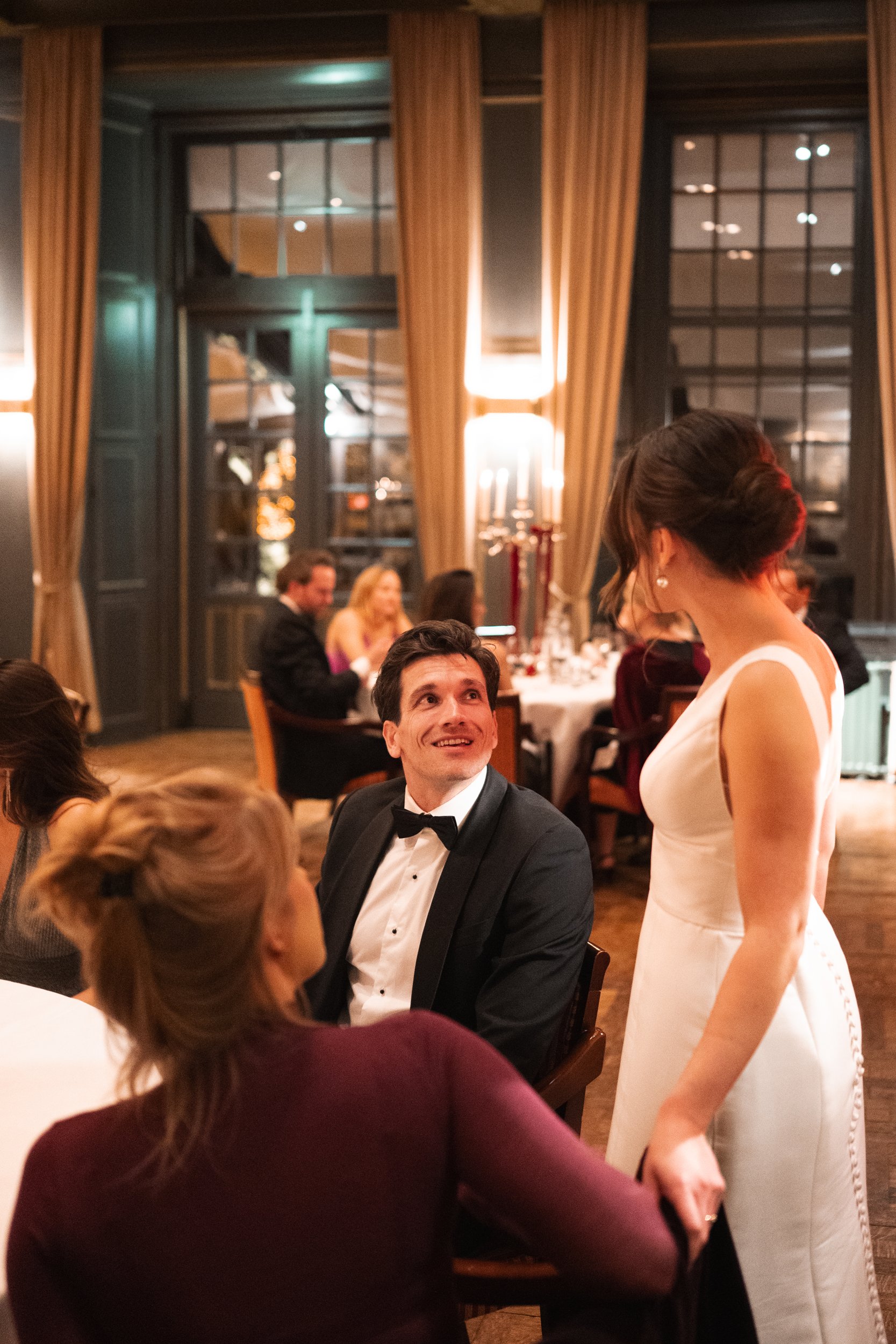 A man in a tuxedo and a woman in a white dress are at a dinner reception, engaging in conversation, surrounded by other guests in an elegant, warmly lit restaurant with large windows and drapes.