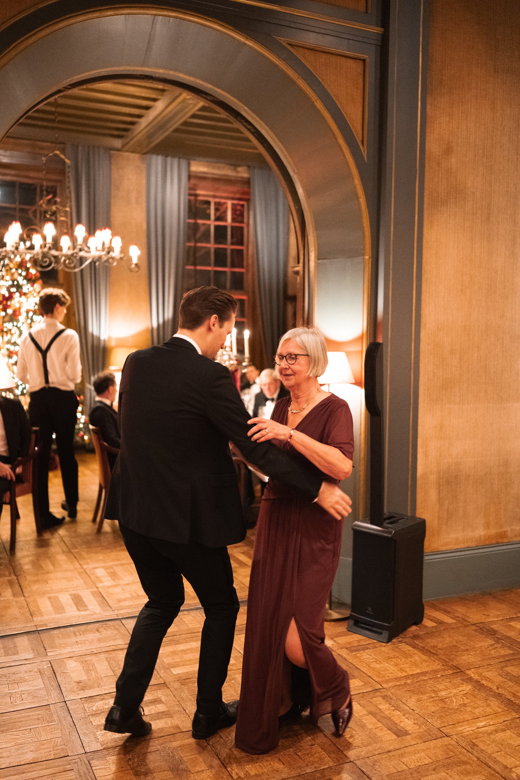 A young man in a black suit and woman in a burgundy dress dancing together in a warmly lit room decorated for Christmas.