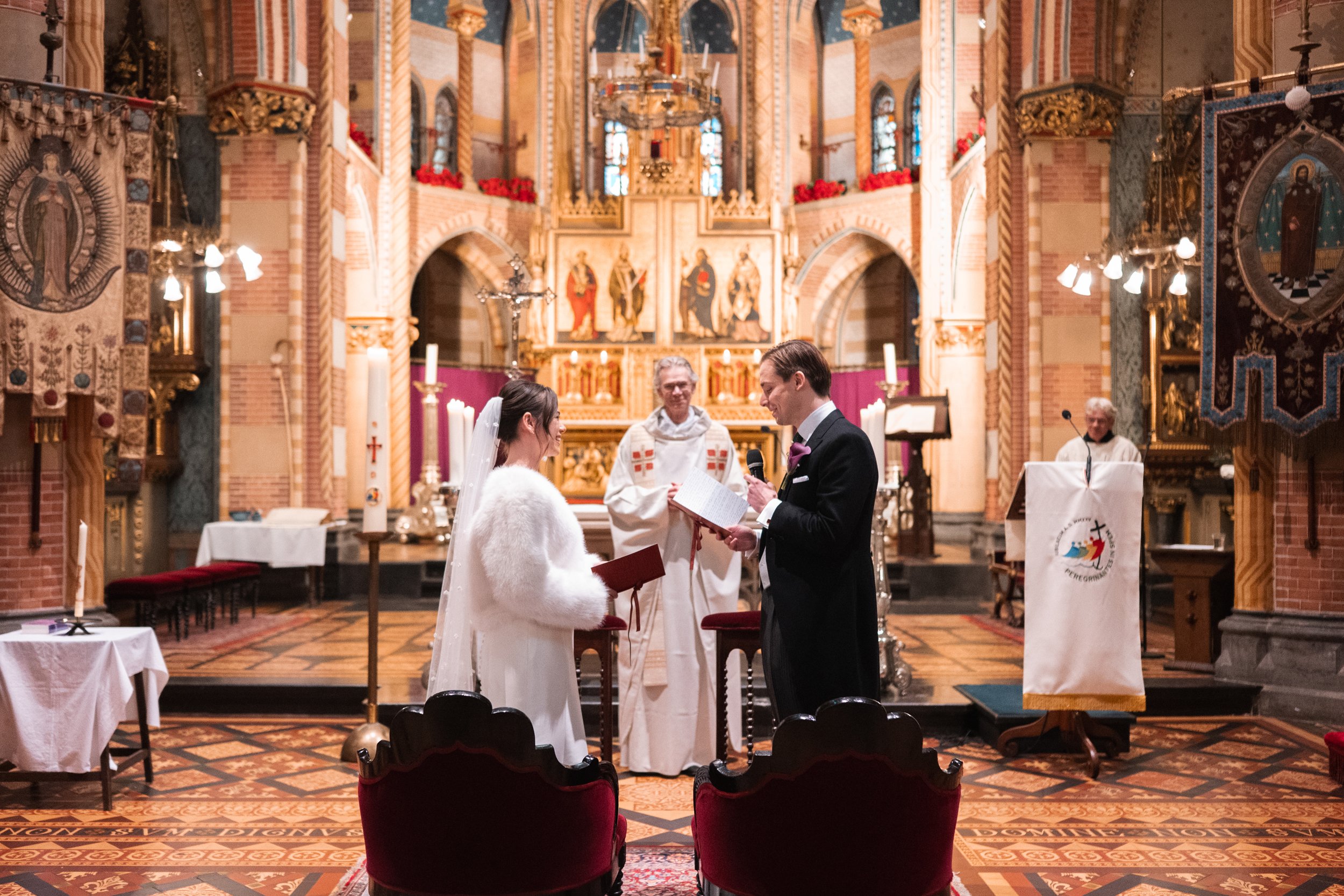 A bride and groom exchanging vows during a wedding ceremony in a church, with an officiant standing behind them and a priest in the background. The church has ornate decorations, candles, and religious icons.