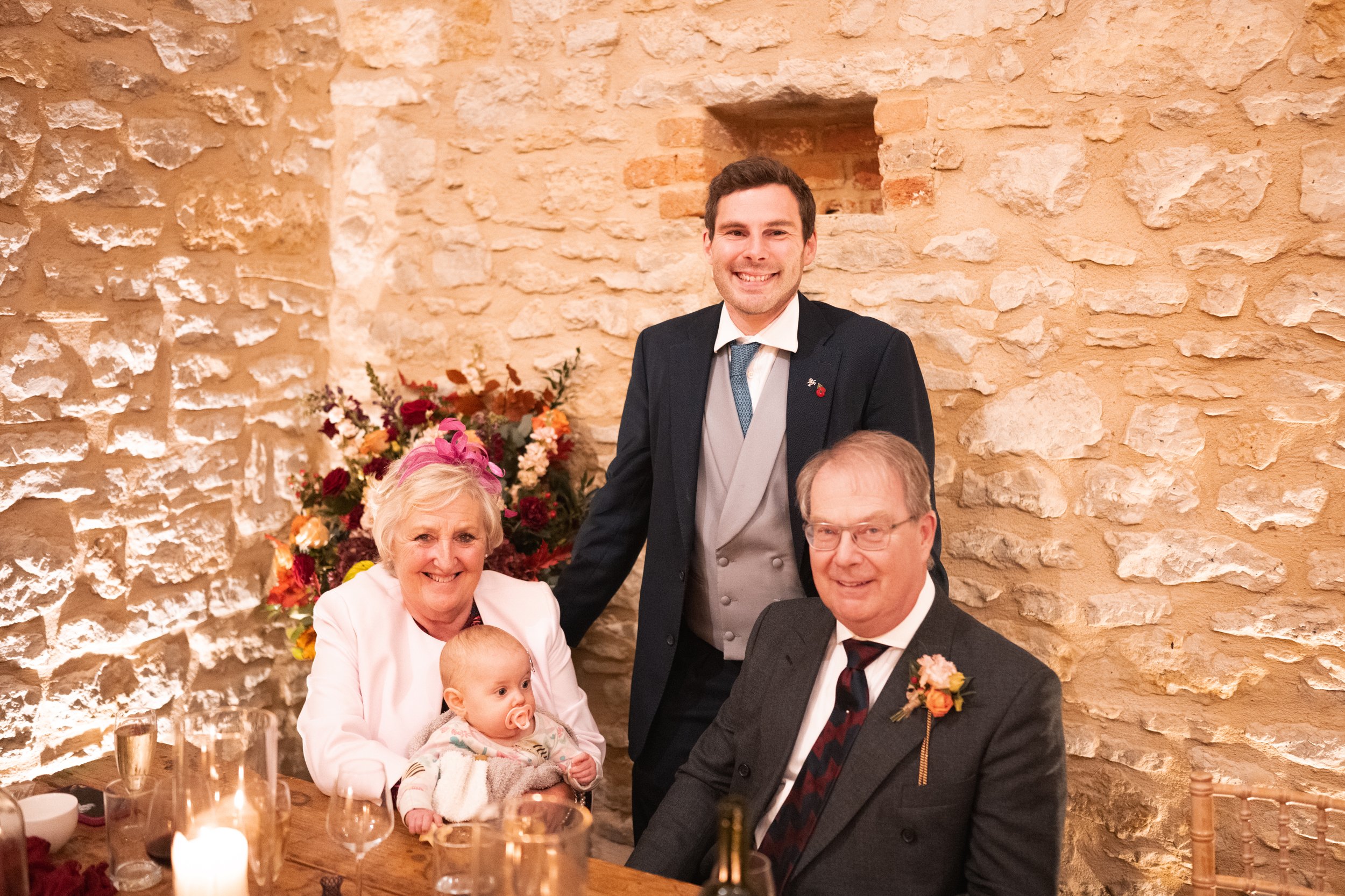 A family celebrating at a dinner table with a man standing behind two seated women with a baby, set against a brick wall and decorated with flowers and candles.
