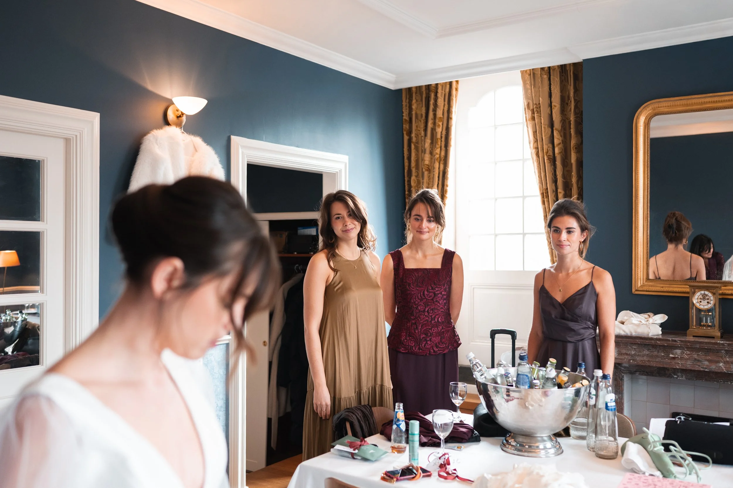 Three women standing behind a table with drinks and decorations, preparing for a special event, while another woman in the foreground adjusts her dress, in a well-lit room with large window and elegant decor.