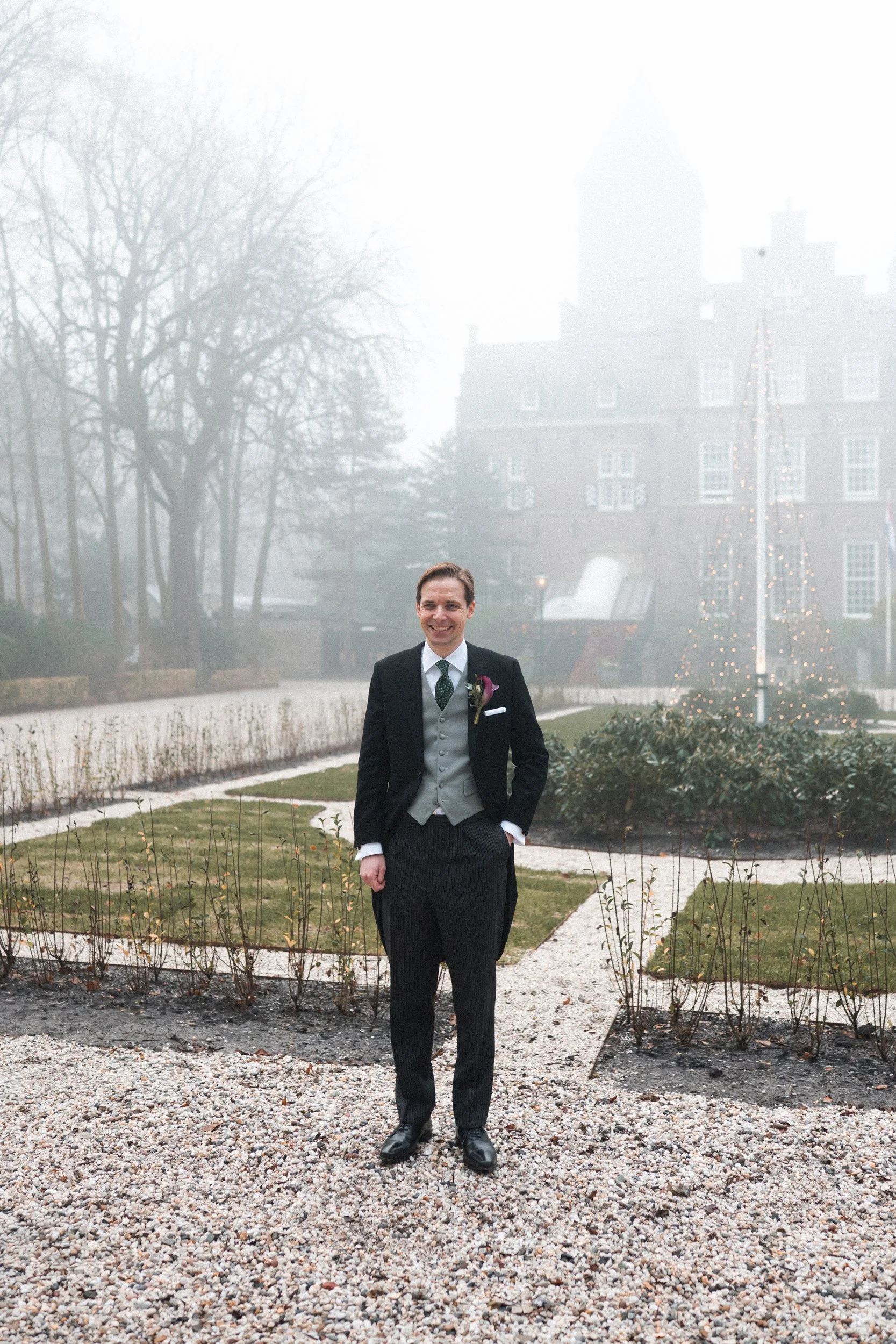 A man dressed in a tuxedo standing outdoors on a foggy day with a historic building and Christmas tree in the background.