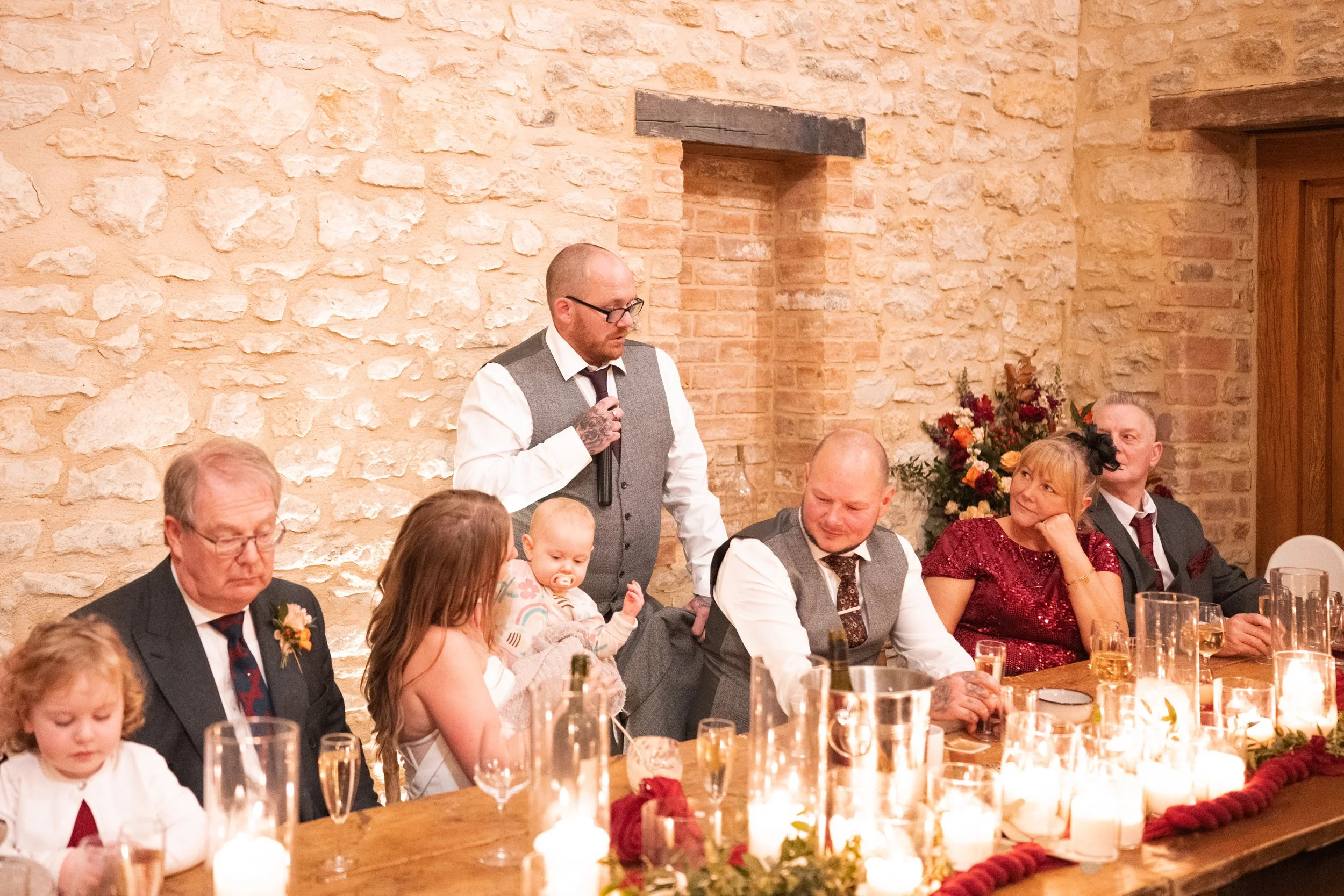 A man standing and speaking into a microphone at a decorated dinner table with several seated adults and children, candles, flowers, and a brick wall background.