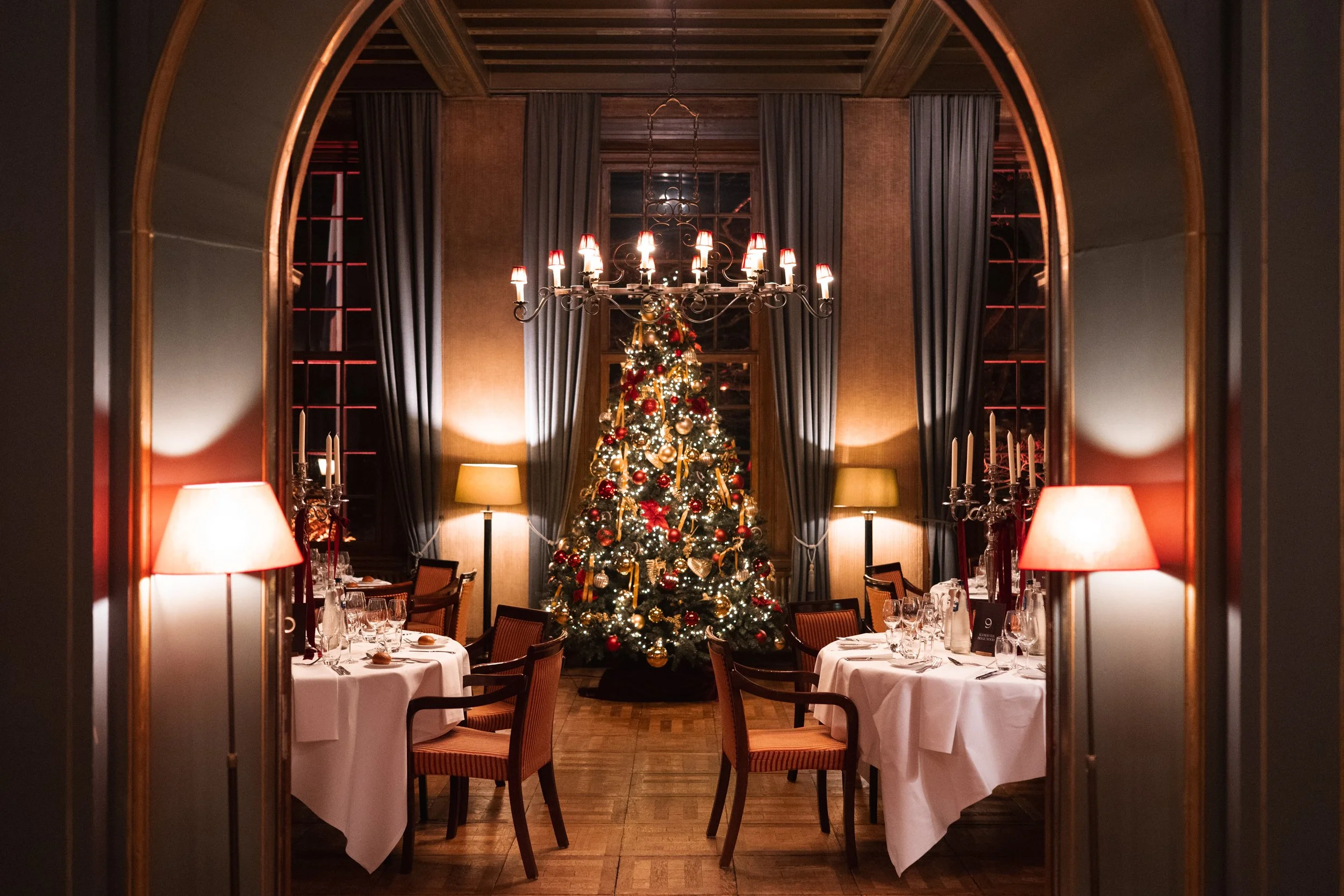 A dining room decorated for Christmas with a lit Christmas tree in the center, surrounded by tables set with white tablecloths, glassware, and candles. The room features wall-mounted lamps, tall curtains, a chandelier, and warm lighting.