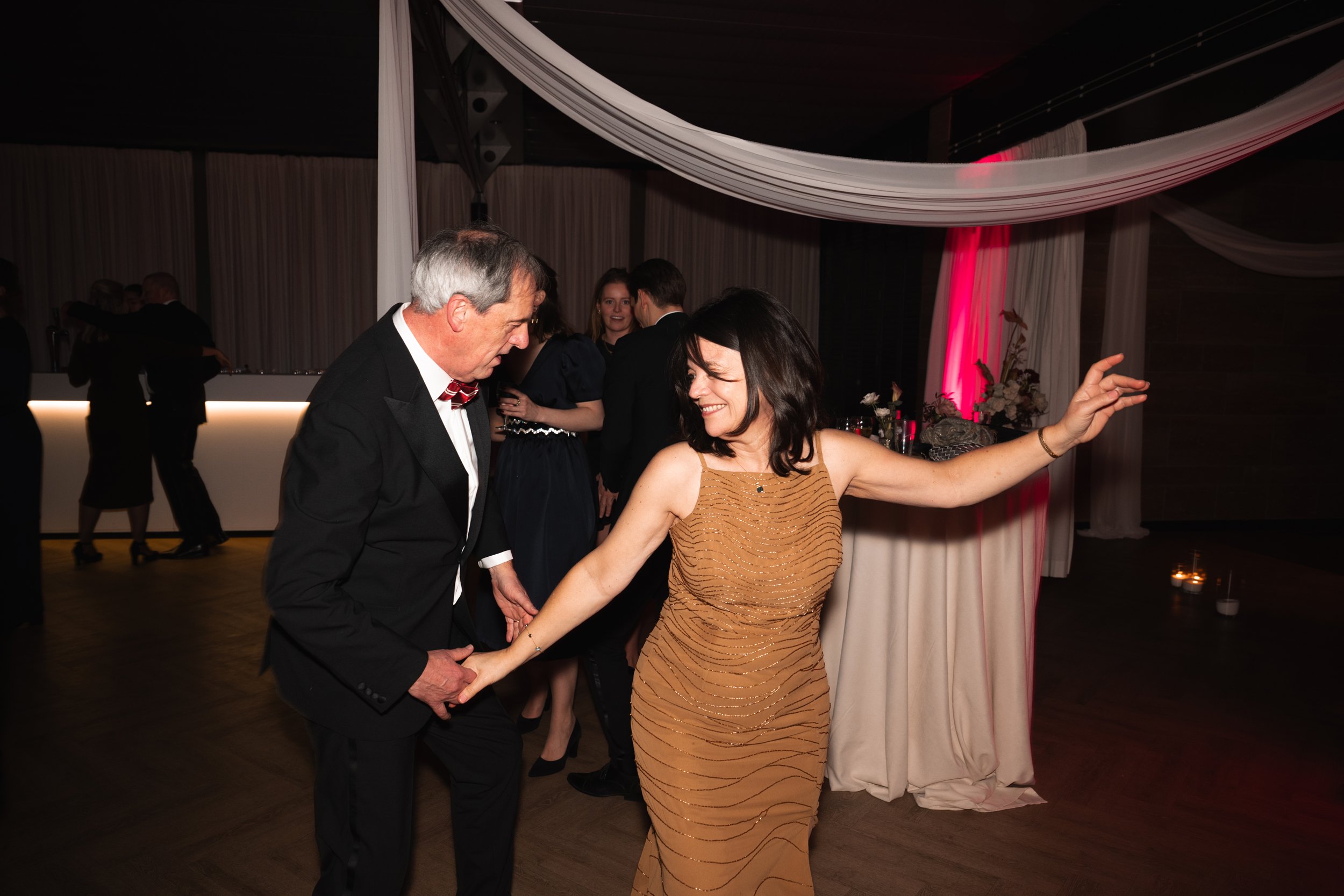 People dancing at a formal event, two women in the foreground holding hands and smiling, other guests in the background, dim lighting and decorative drapes.