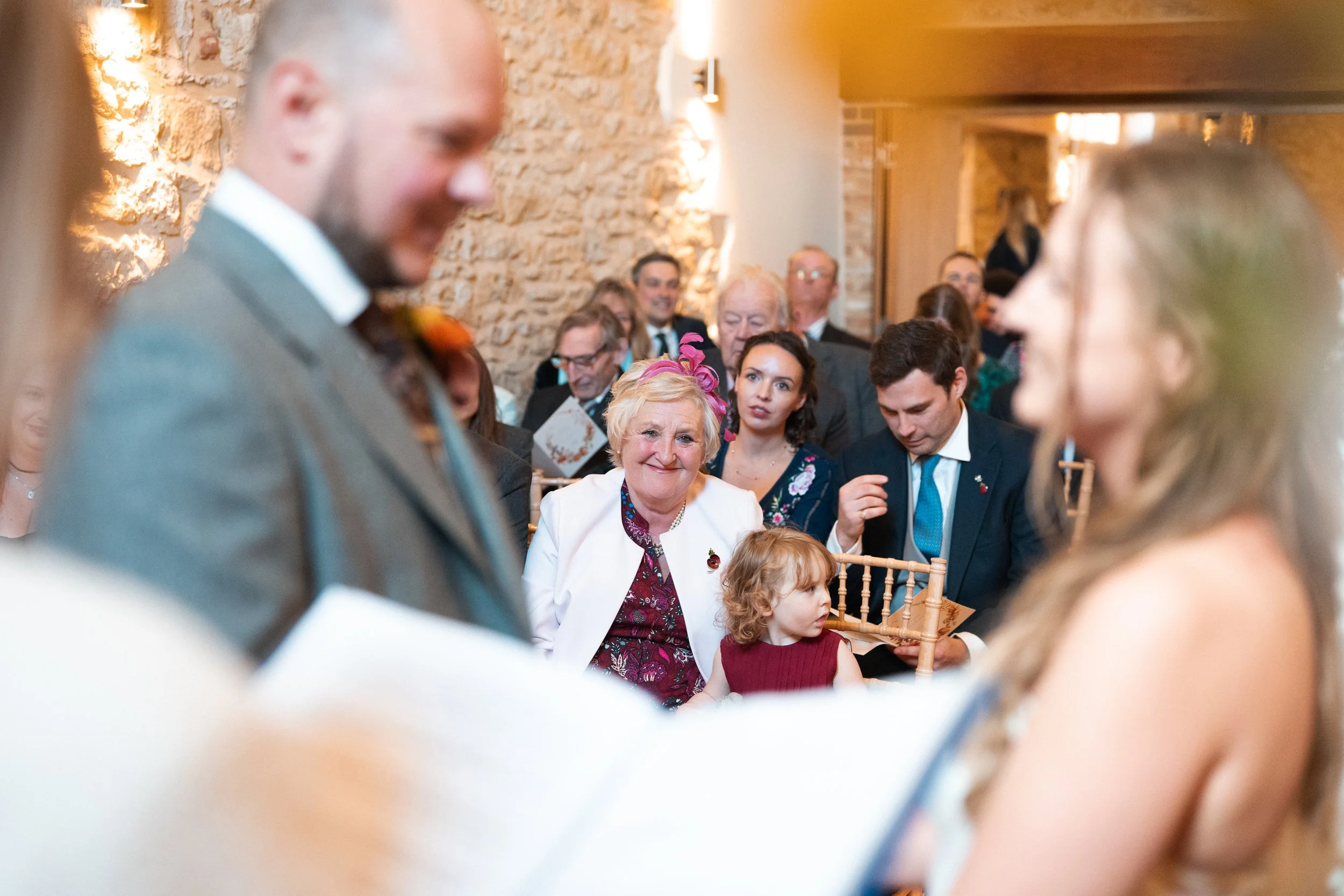 People attending a wedding ceremony, with a focus on an elderly woman smiling at a young couple, surrounded by guests in an indoor venue with exposed brick walls.