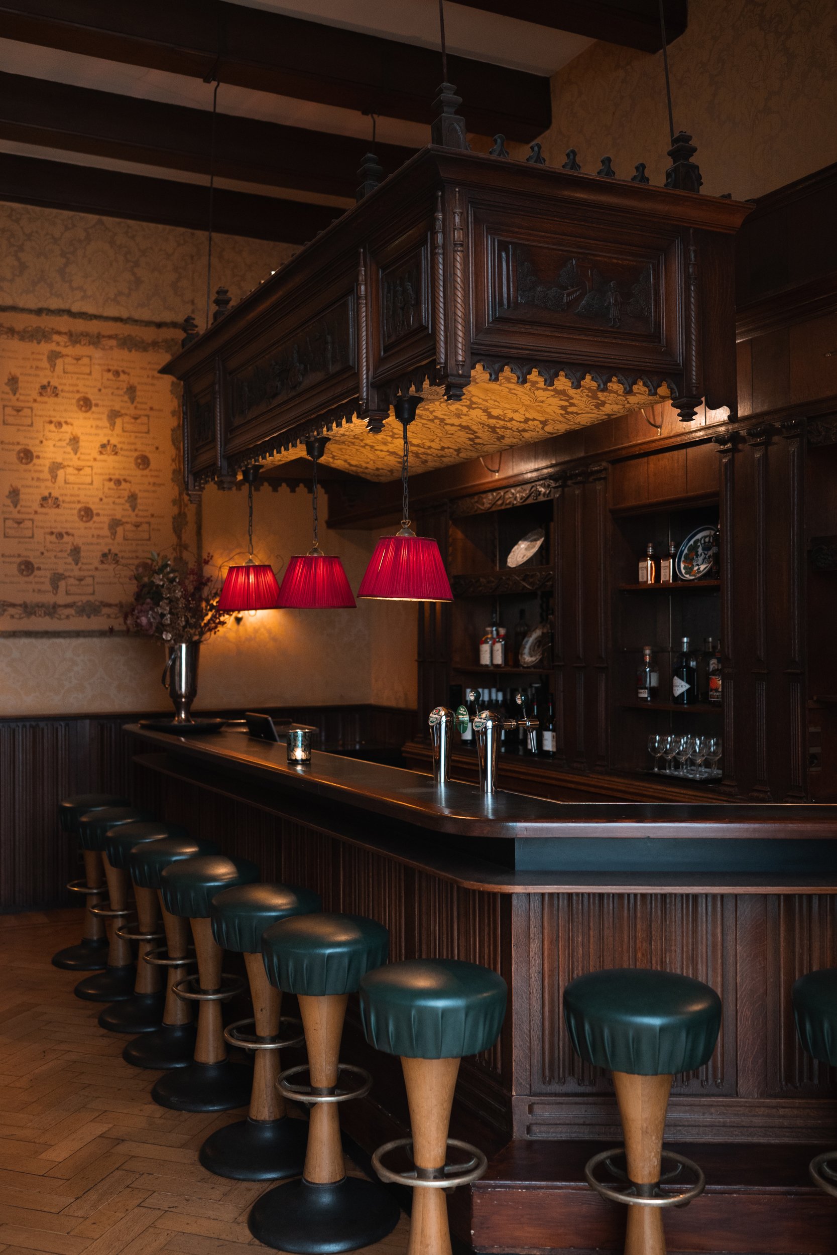 A vintage-style bar with a wooden counter, green upholstered stools, dark wooden shelves holding bottles of alcohol, red lampshades hanging above, and ornate wood carvings.