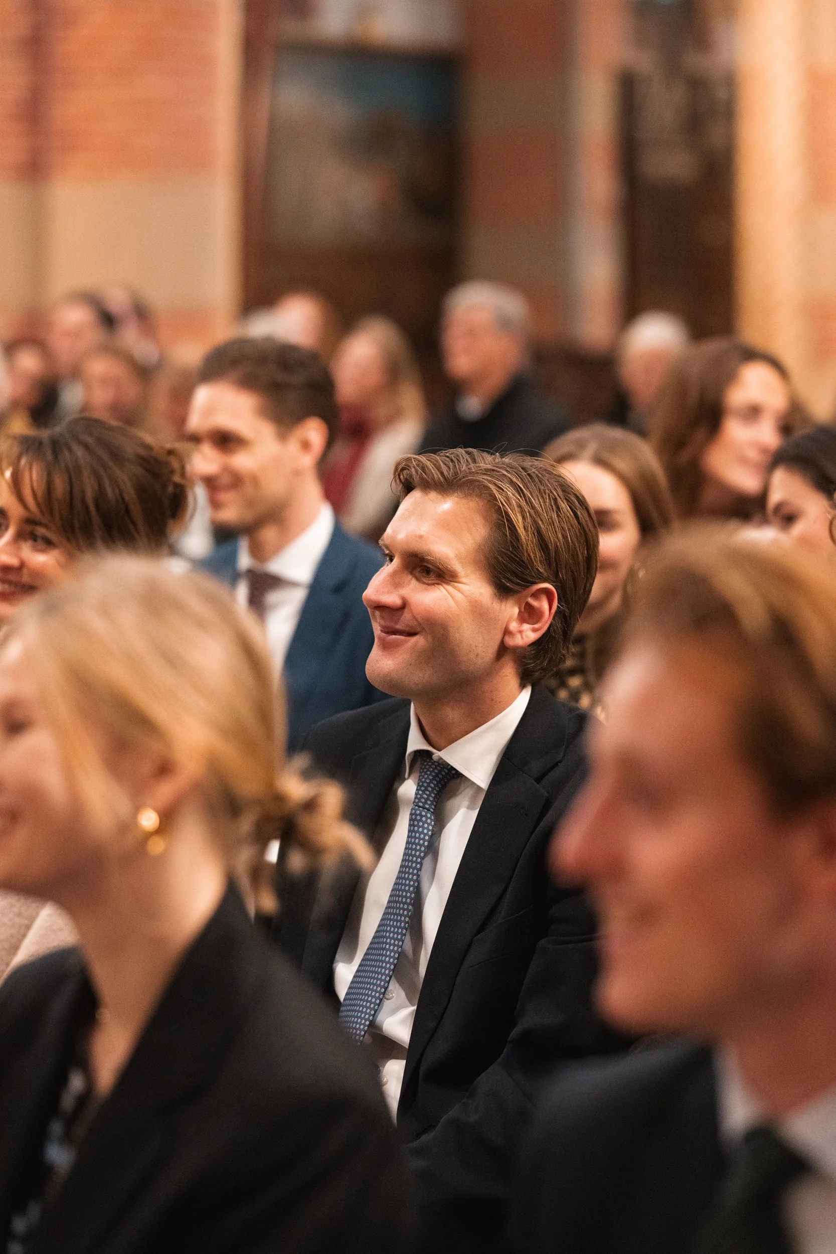A group of well-dressed people attending a formal event, seated and listening attentively, with a man smiling in the center.