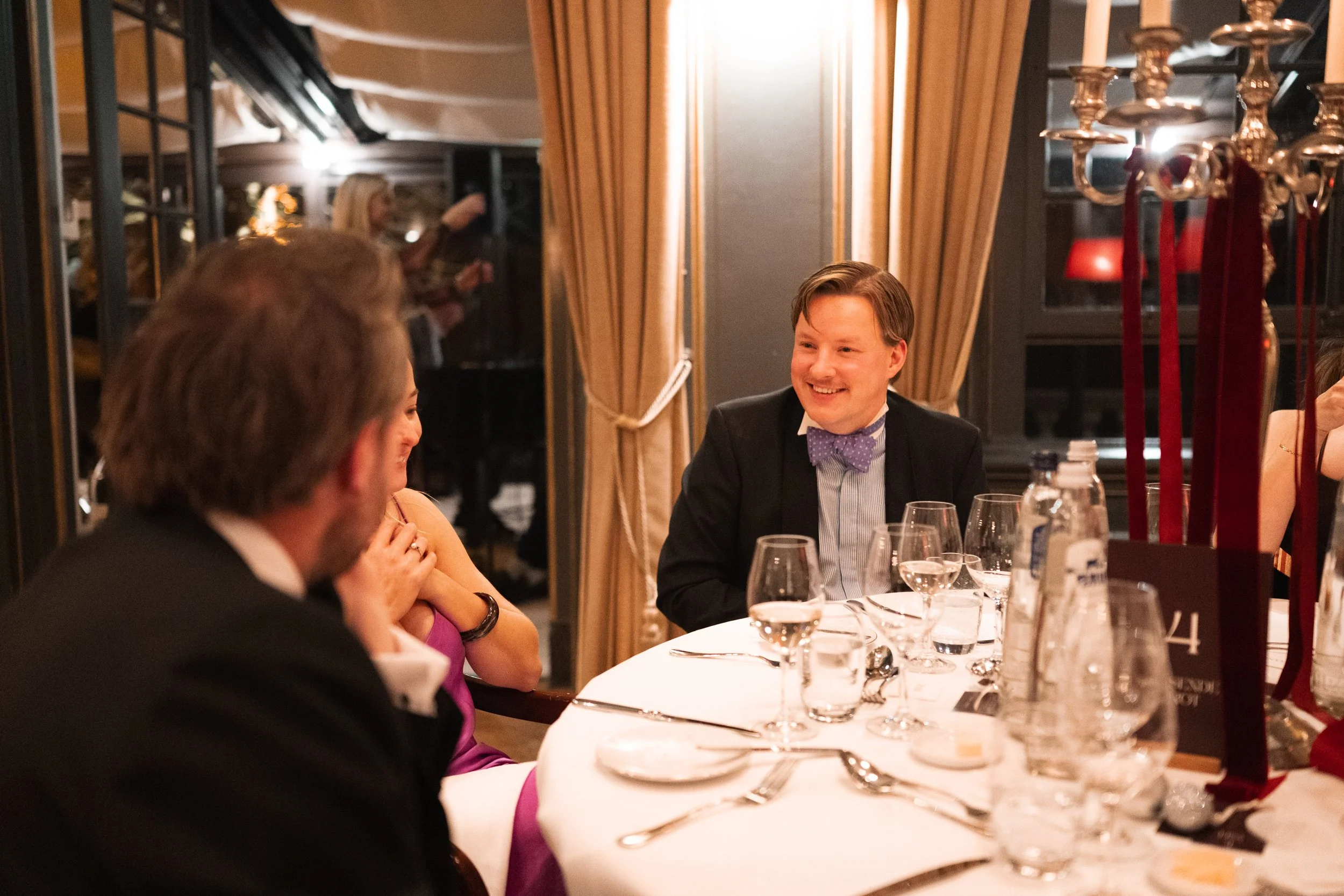 People seated at a dinner table in a formal setting, smiling and talking, with wine glasses and tableware.