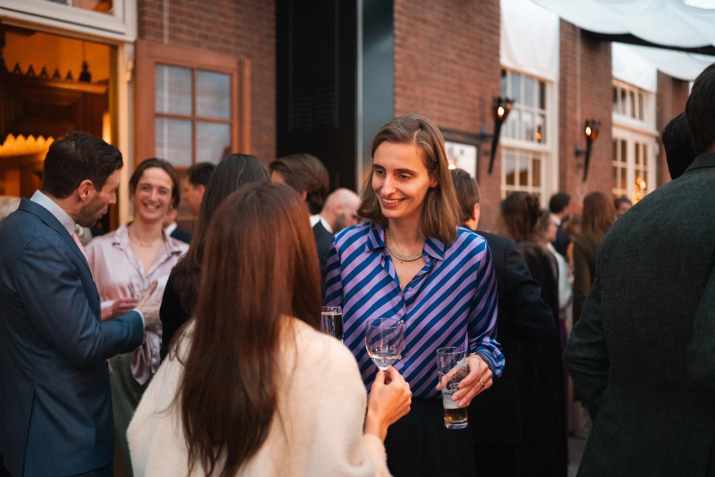 Group of people socializing at a party in a brick-walled venue, some with drinks, engaging in conversations, with warm lighting and large windows in the background.