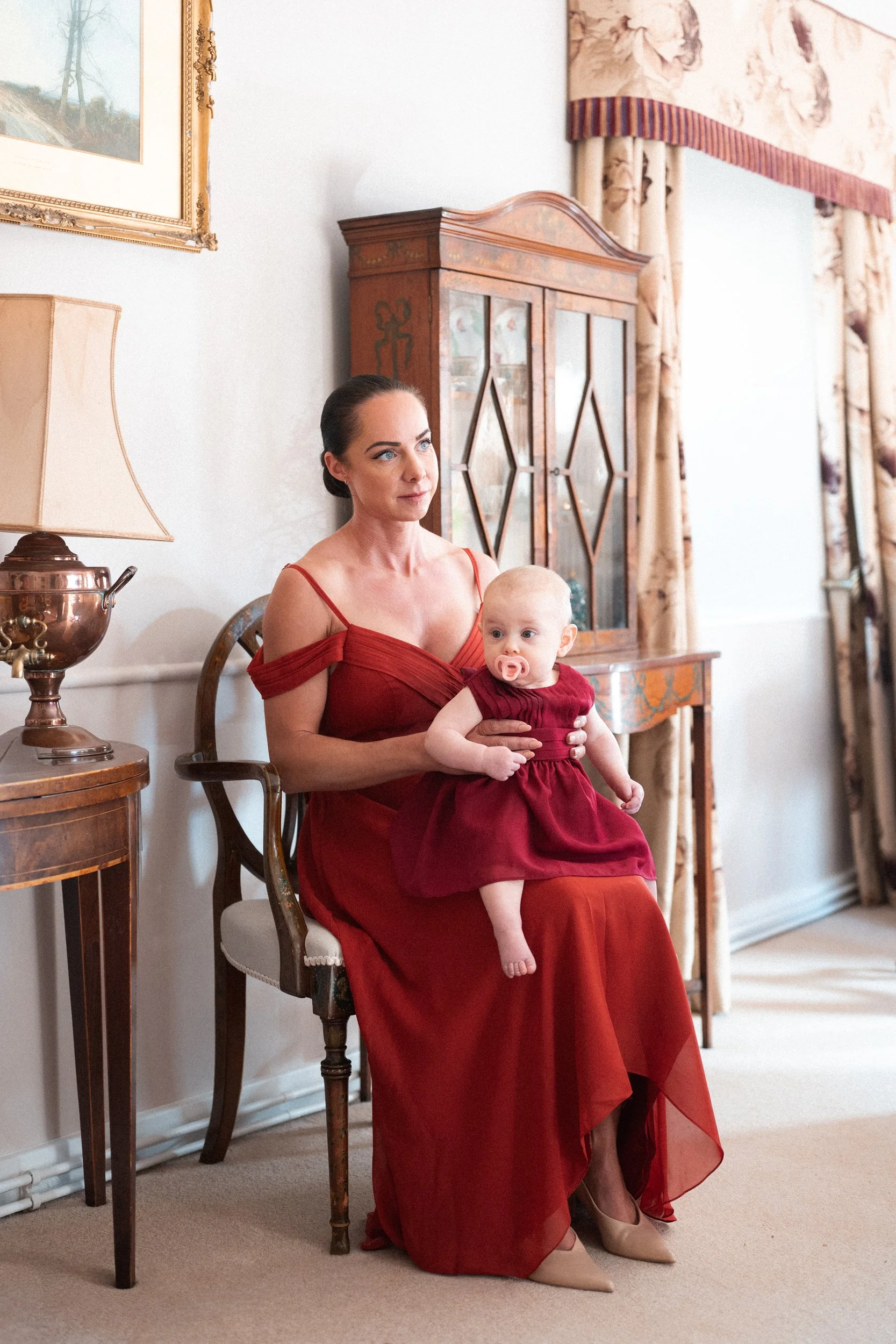 A woman in a red dress sitting on a chair, holding a baby girl in a matching red dress, in a living room with vintage furniture and curtains.