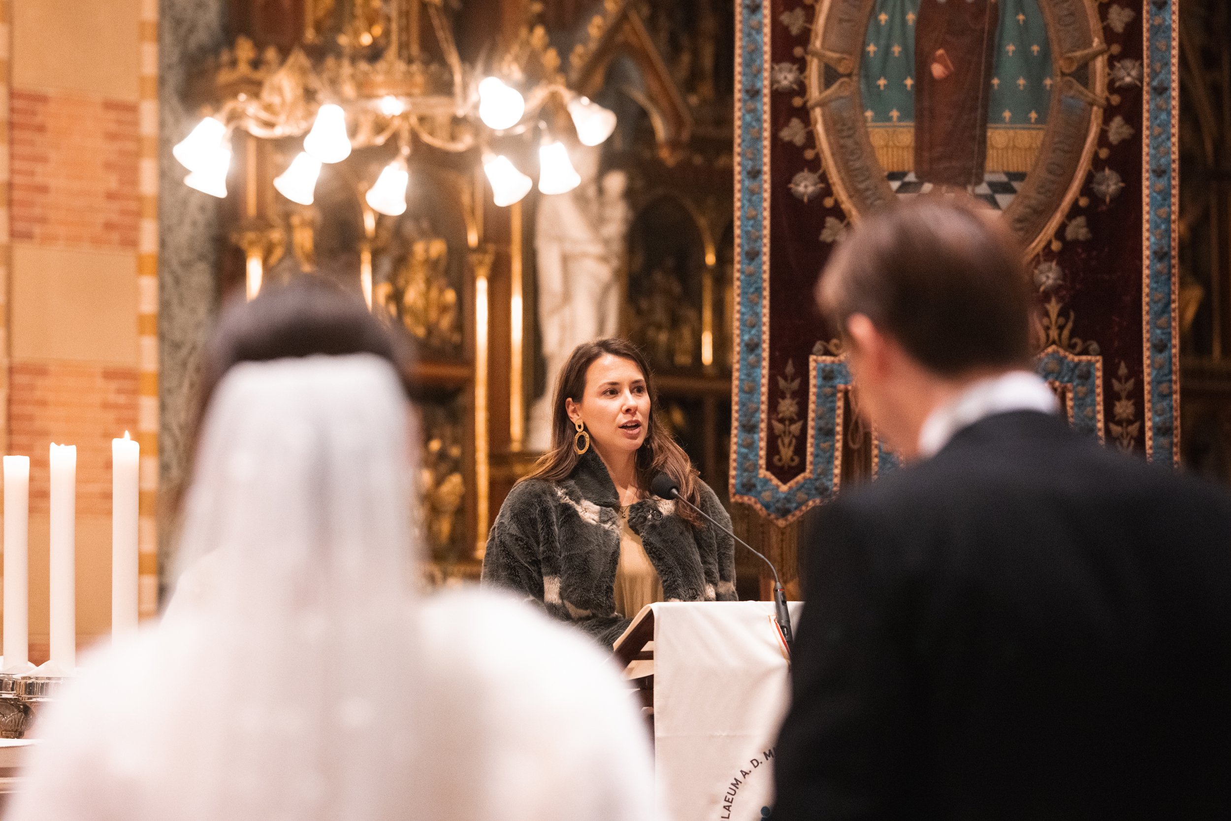 A woman is speaking at a podium inside a church, with a man and a woman seated in the foreground. The church has ornate gold decorations, a large banner, and overhead lighting.