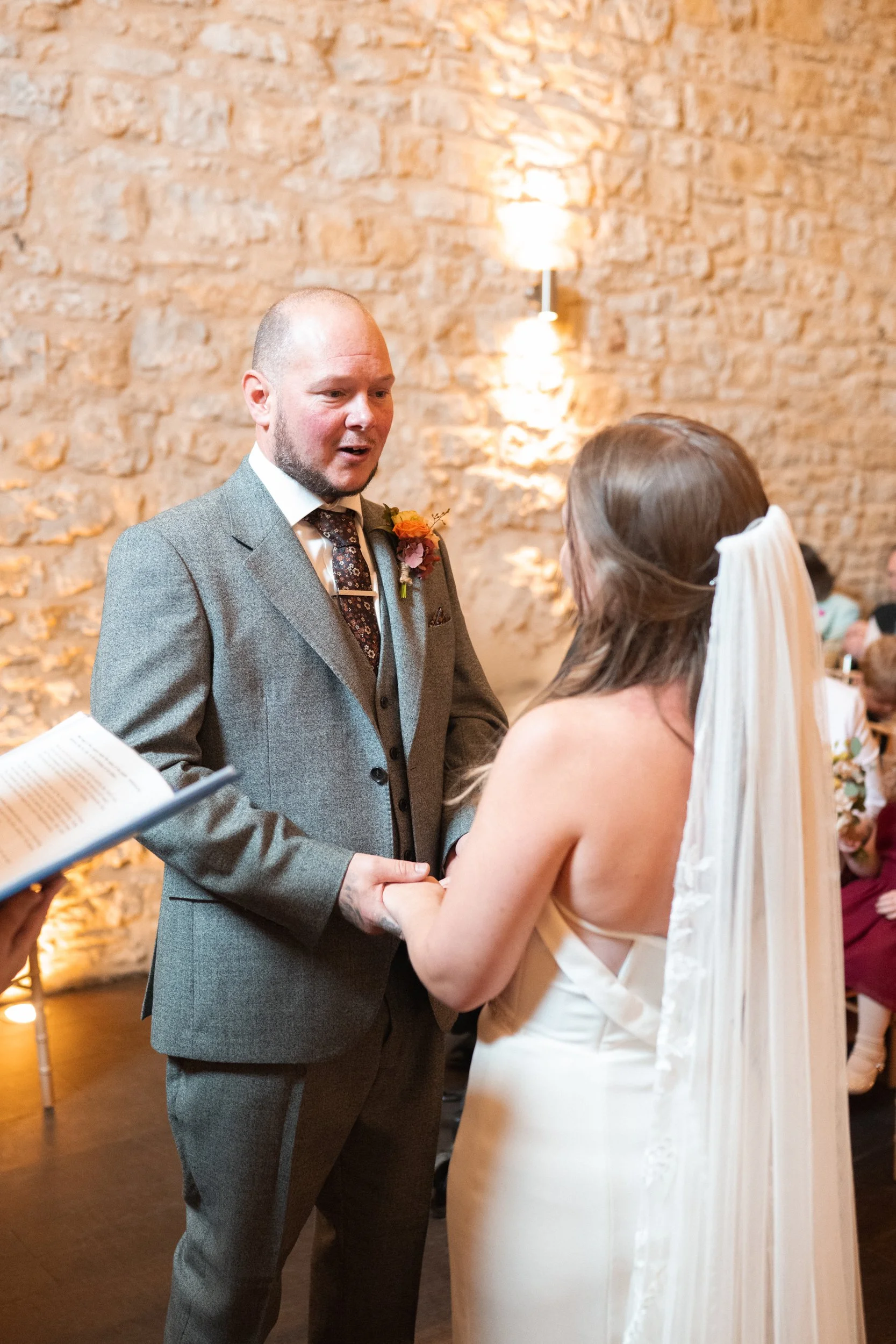 A man in a gray suit and a woman in a white wedding dress hold hands during a wedding ceremony indoors with a brick wall background.
