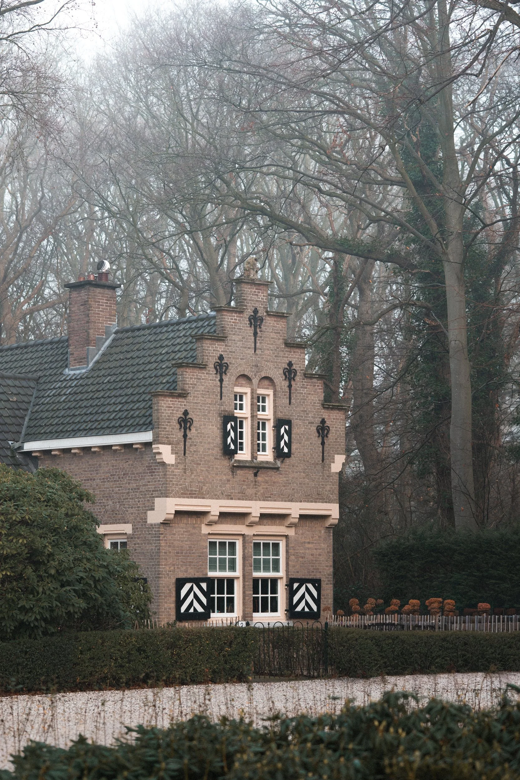 A small brick house with black and white shutters and decorative black ironwork, situated in a wooded area with tall, leafless trees in the background.