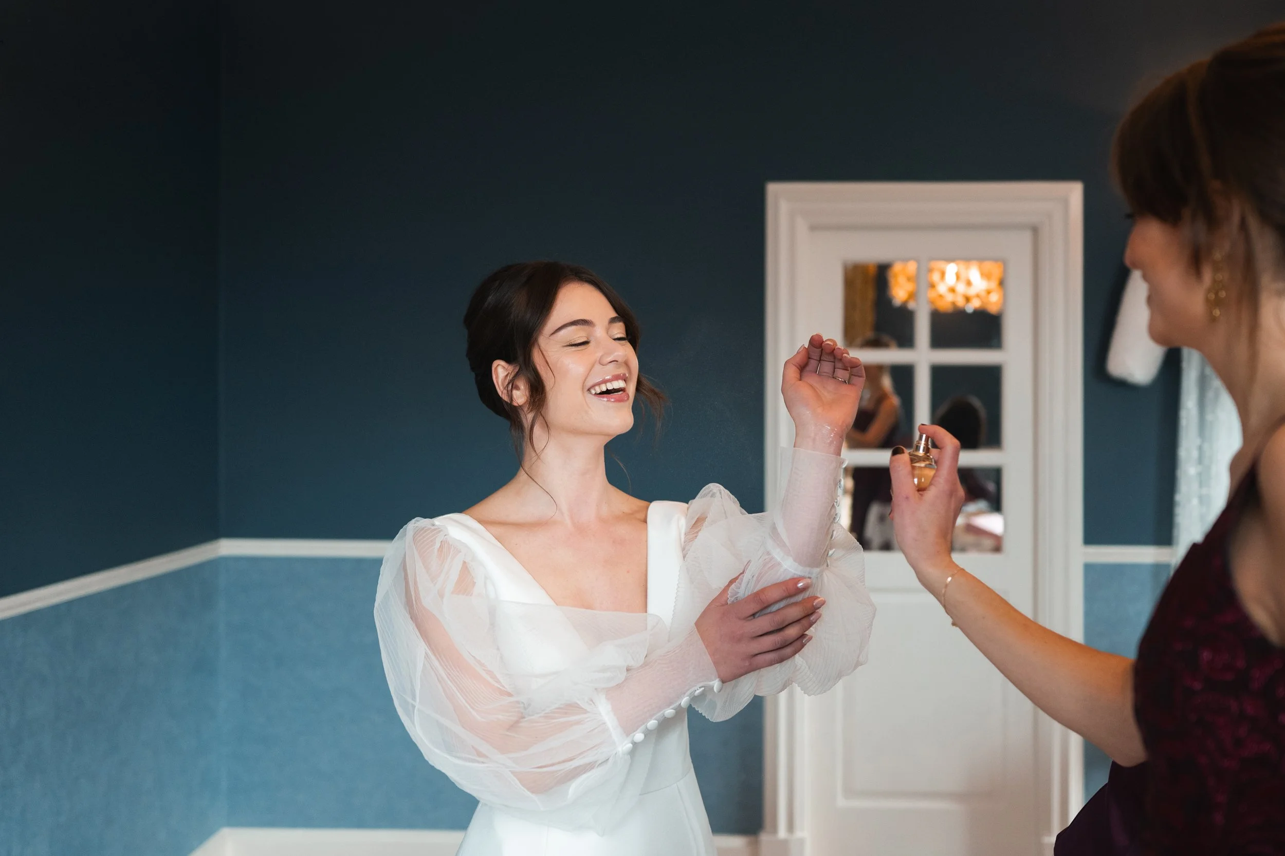 Bride smiling with eyes closed, holding a perfume bottle in her right hand, in a room with blue walls, a mirror, and a woman in a dark dress.