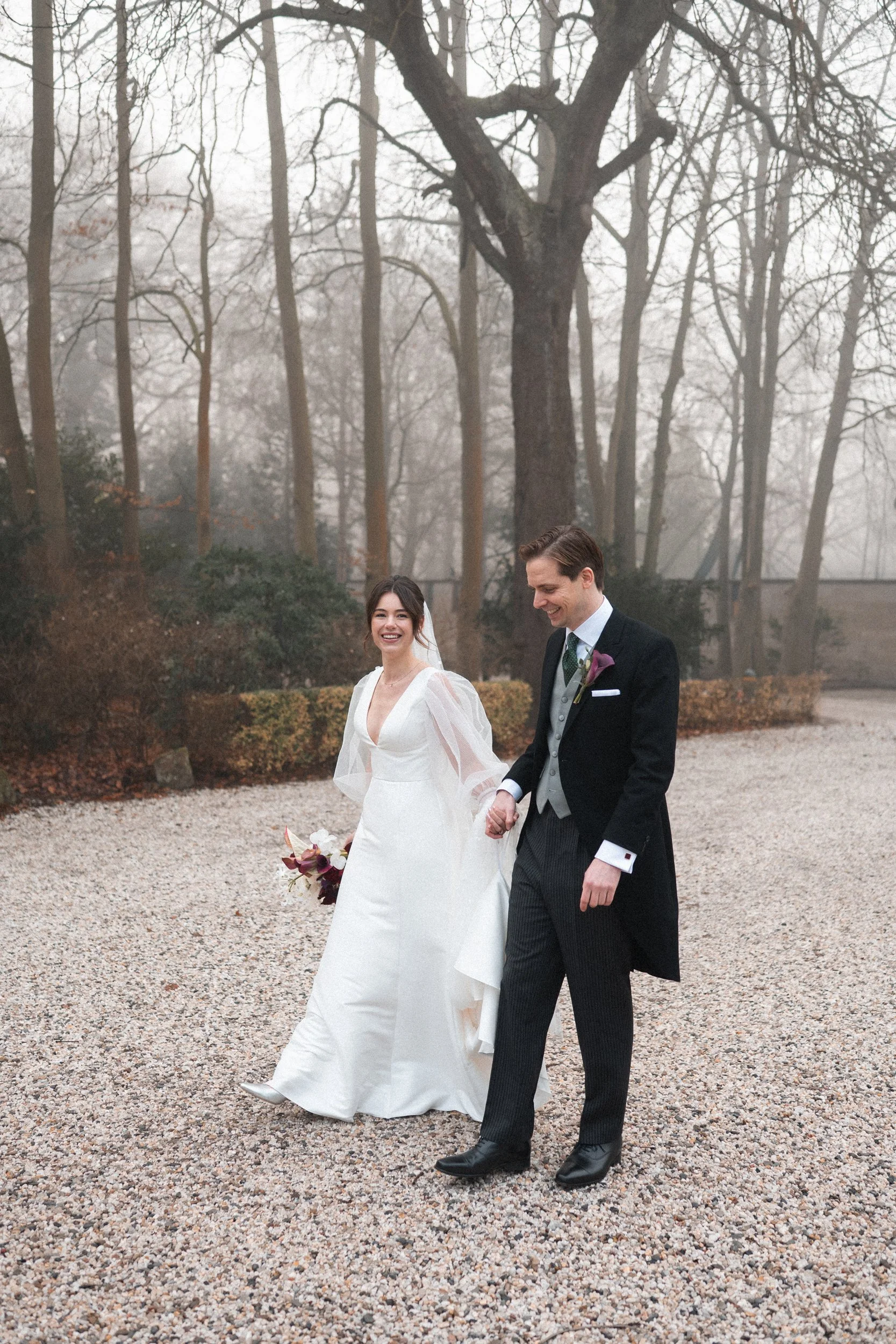 A bride and groom walking hand in hand outdoors on a gravel path with foggy trees in the background during winter.