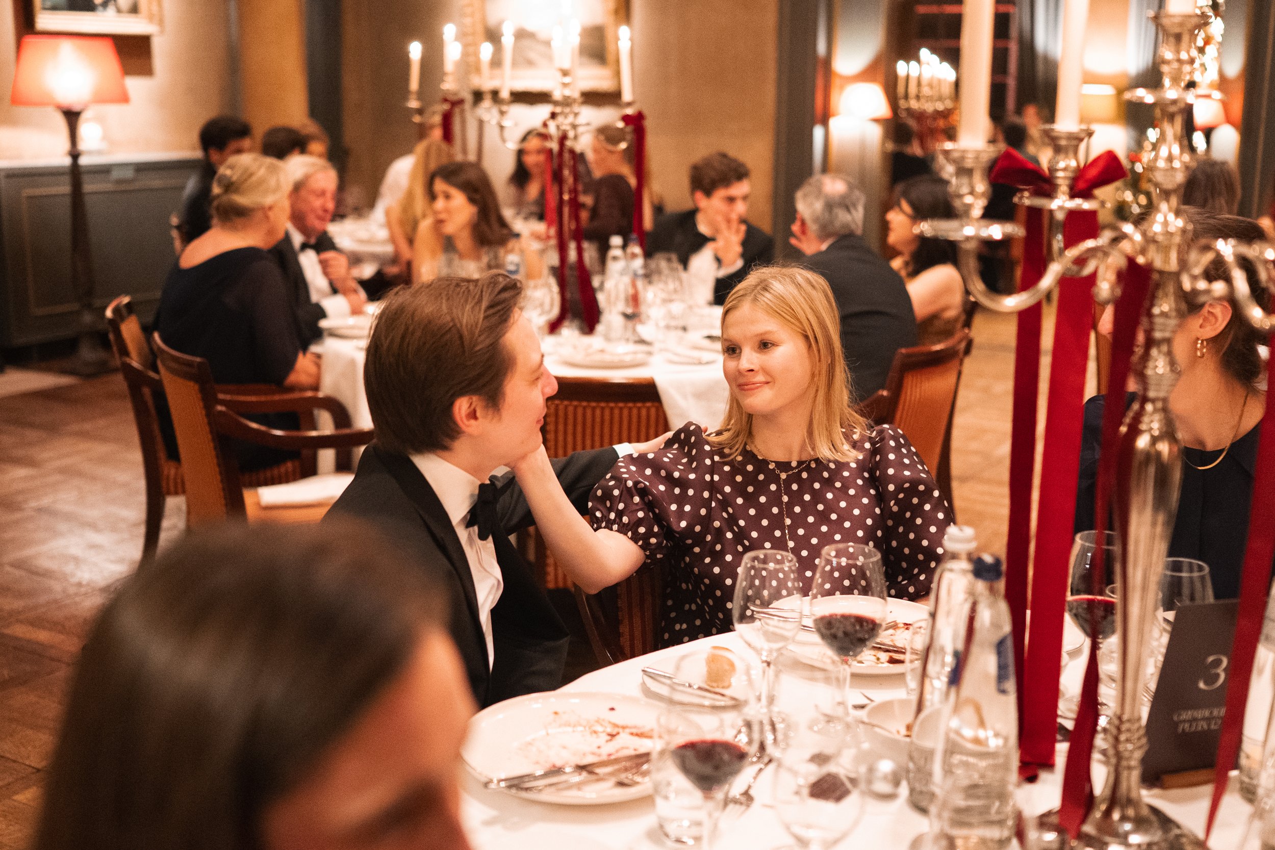 A woman with blonde hair in a polka dot dress gently touches the face of a man in a tuxedo at a formal dinner event. They are seated at a table with wine glasses and dinner plates, and the background shows other guests engaged in conversation in a wa