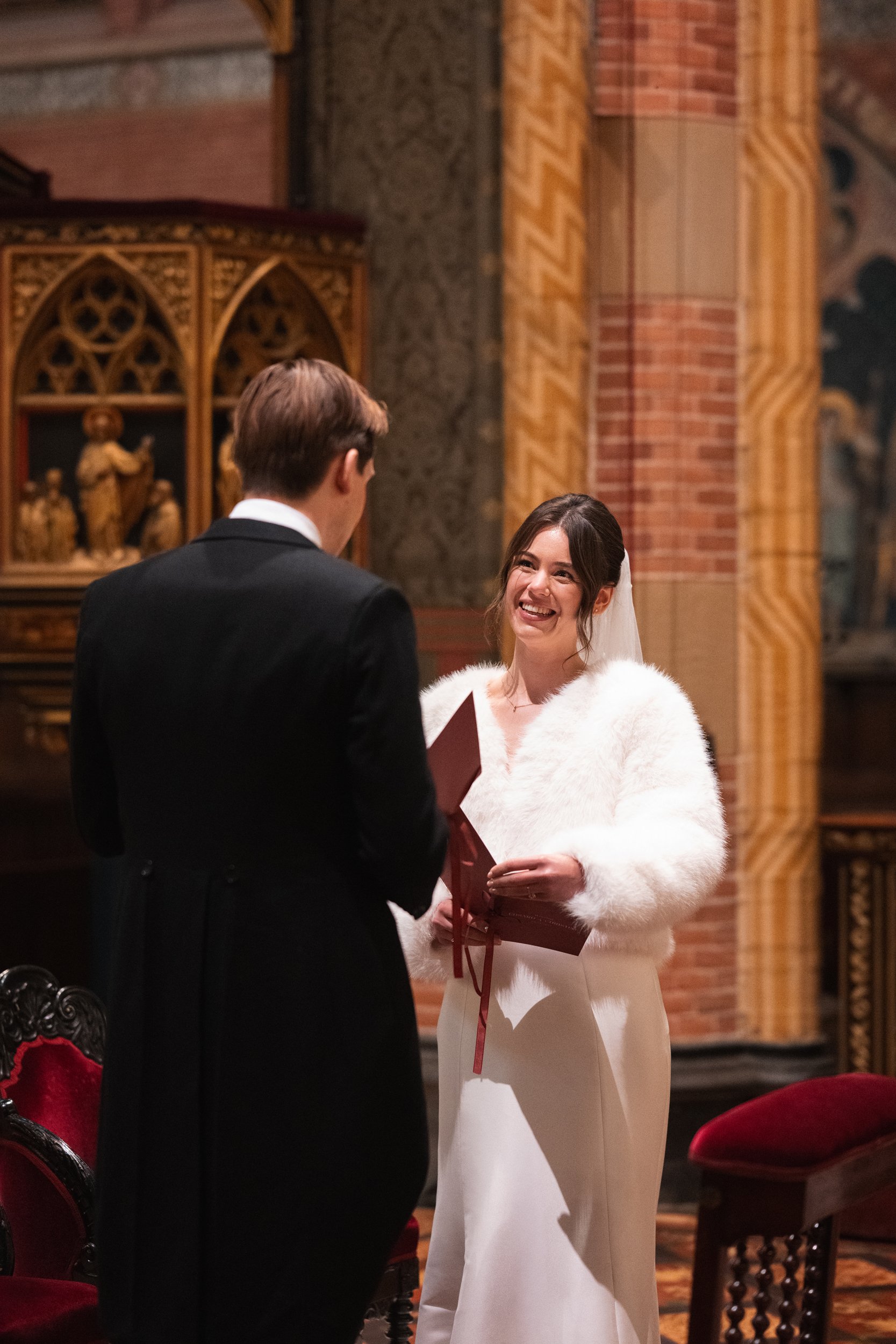 A bride and groom exchanging vows during their wedding ceremony inside a church, with the bride smiling and holding a box, and the groom facing her.