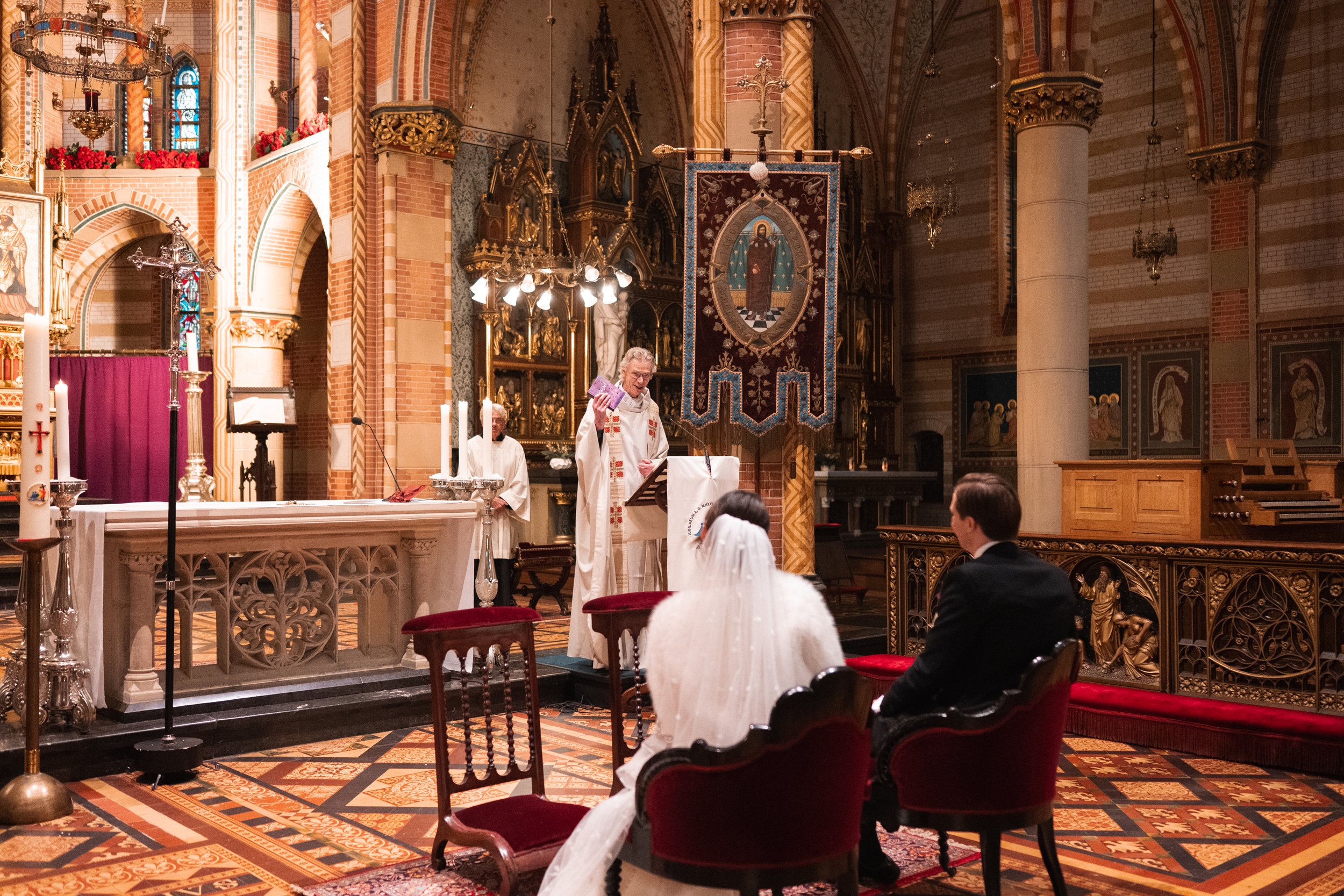 A wedding ceremony taking place inside a church with ornate decorations, stained glass windows, and an altar. The bride and groom are seated facing the priest, who is conducting the ceremony. The priest is holding a booklet, and there are candles and