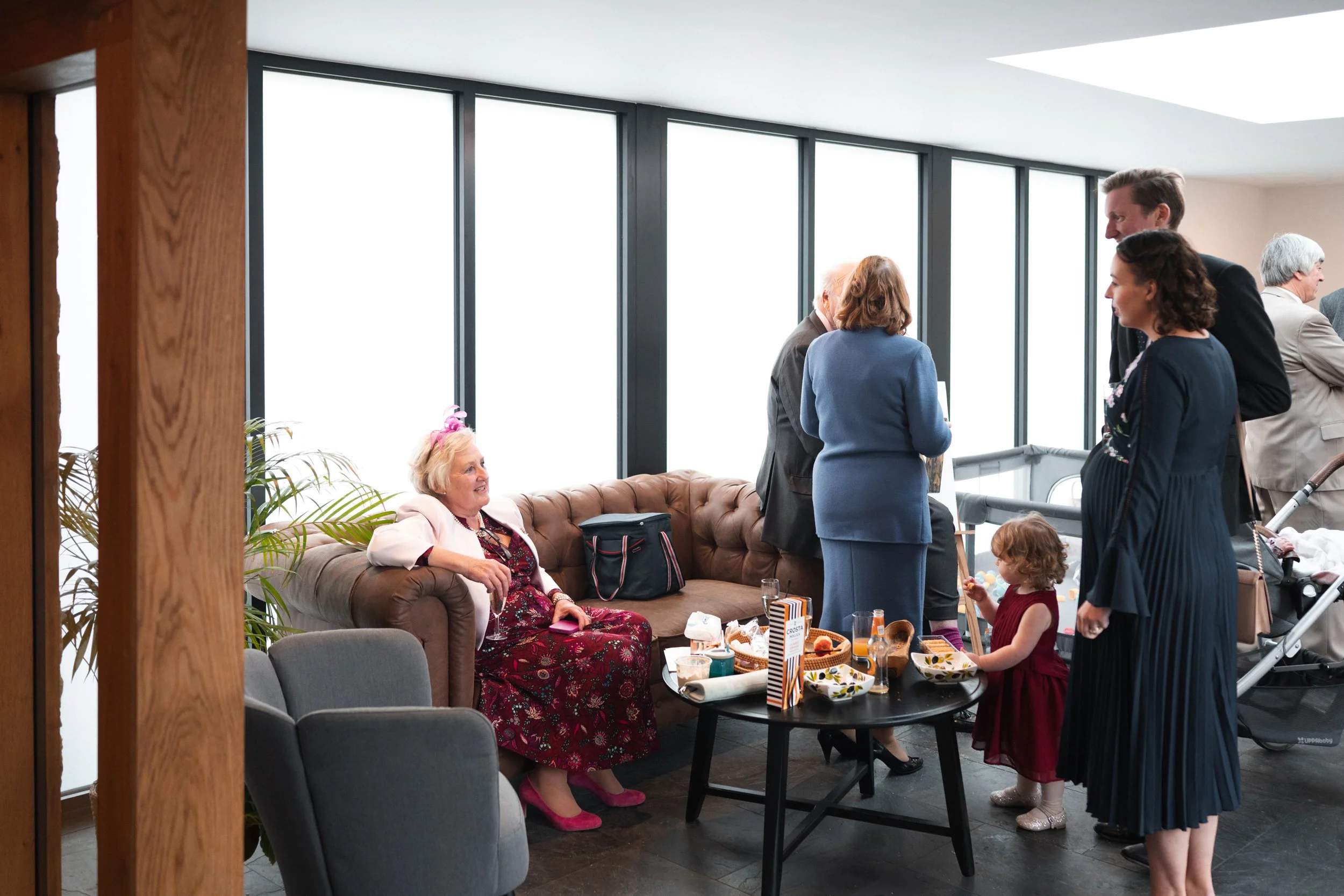 A group of people enjoying a gathering in a bright room with large windows. A woman in a floral dress and pink shoes sits on a brown couch. A young girl in a red dress stands near a table with snacks and drinks. Several adults are standing and talkin