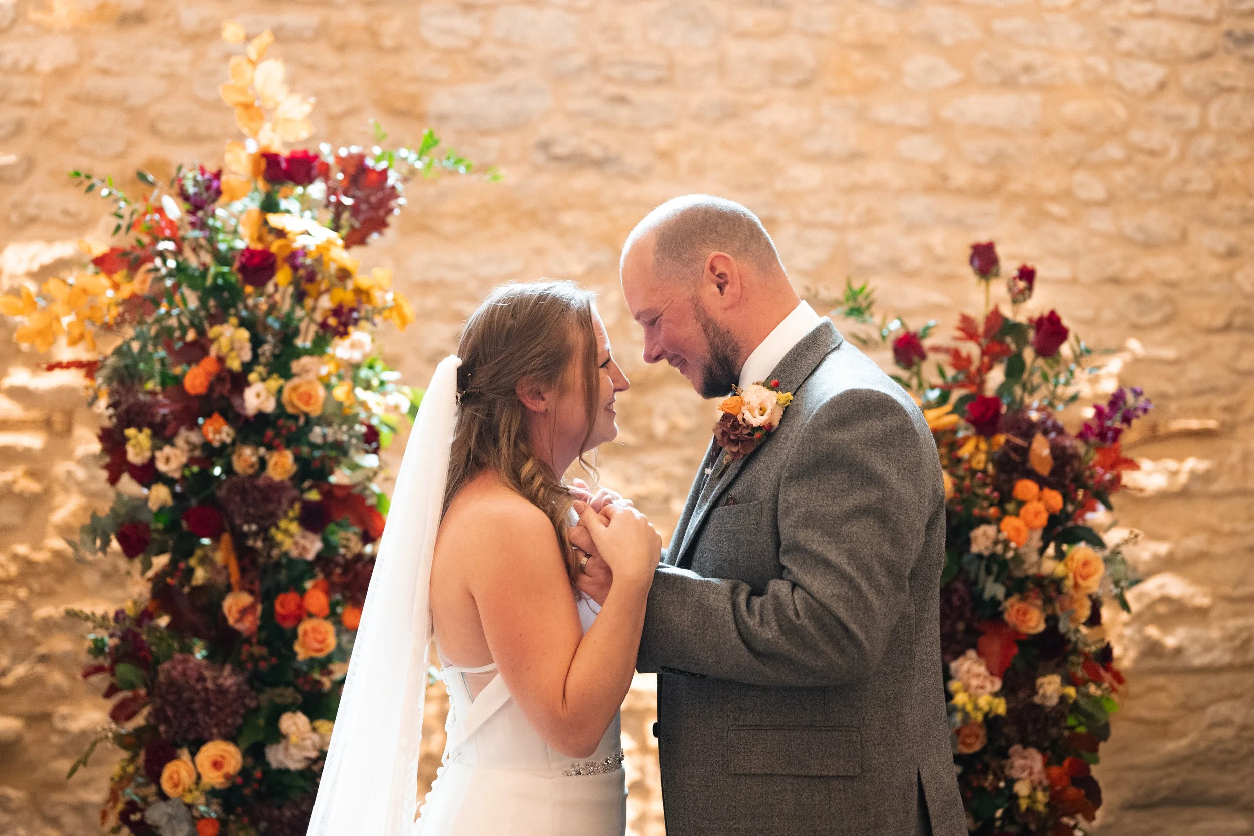 Bride and groom standing close, smiling, and touching foreheads during their wedding ceremony, with colorful floral arrangements in the background and a stone wall.