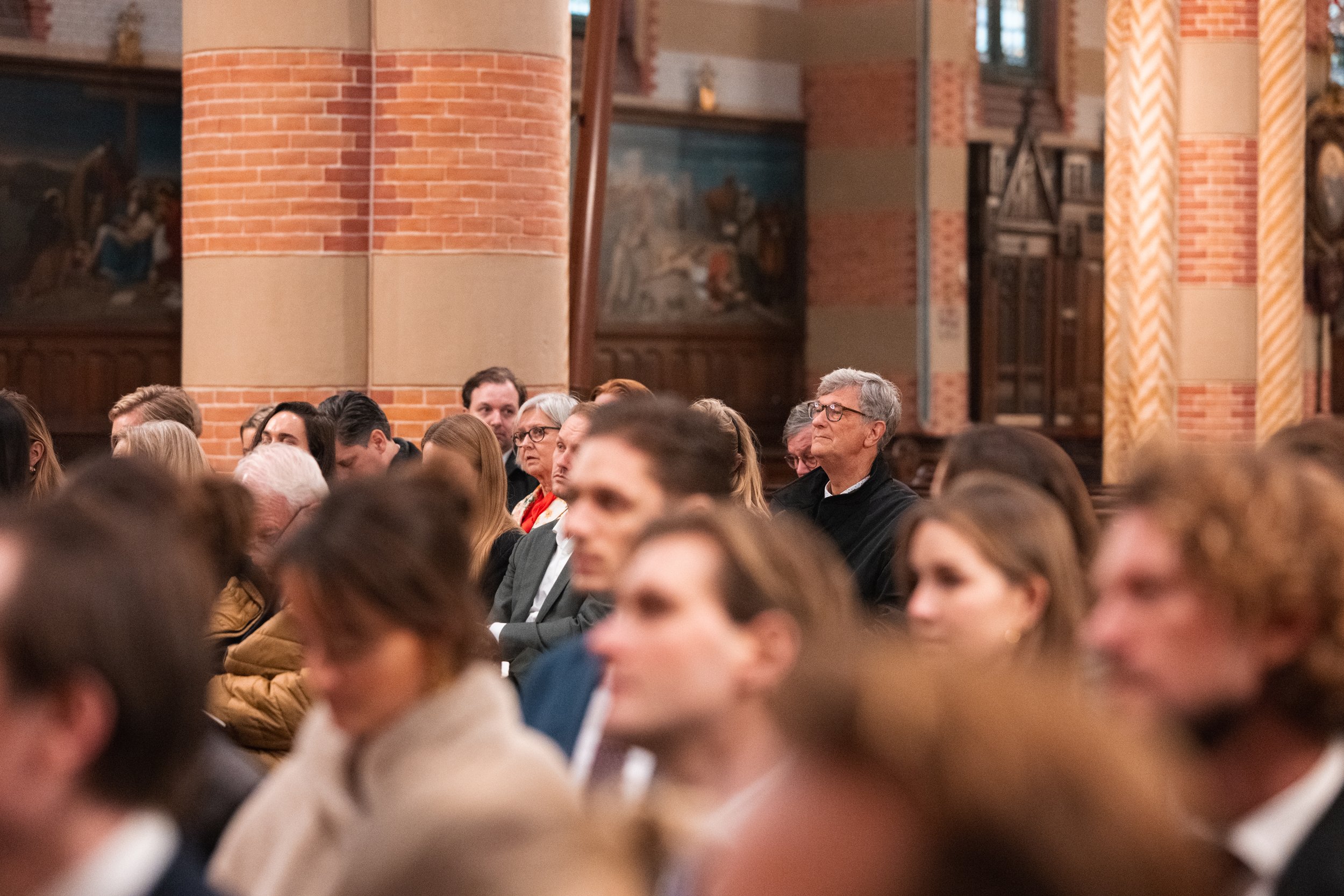 People attending a formal event inside a church, seated in rows facing forward, with brick columns and religious artwork in the background.