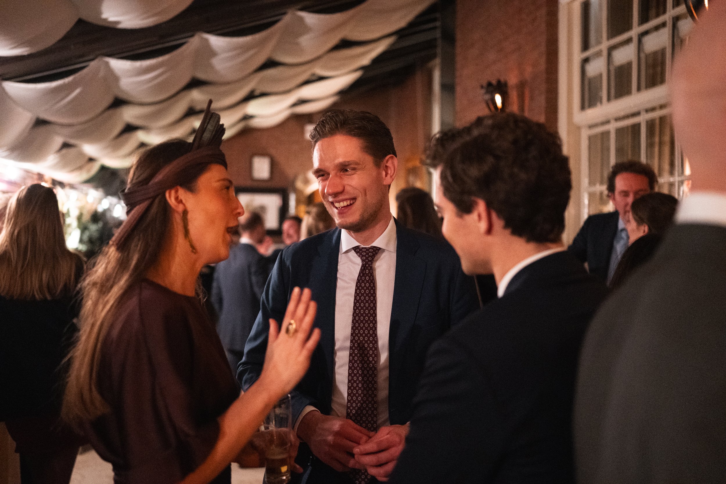 People socializing at a party, engaging in conversation and smiling in a warmly lit indoor setting with decorative ceiling drapes and window.