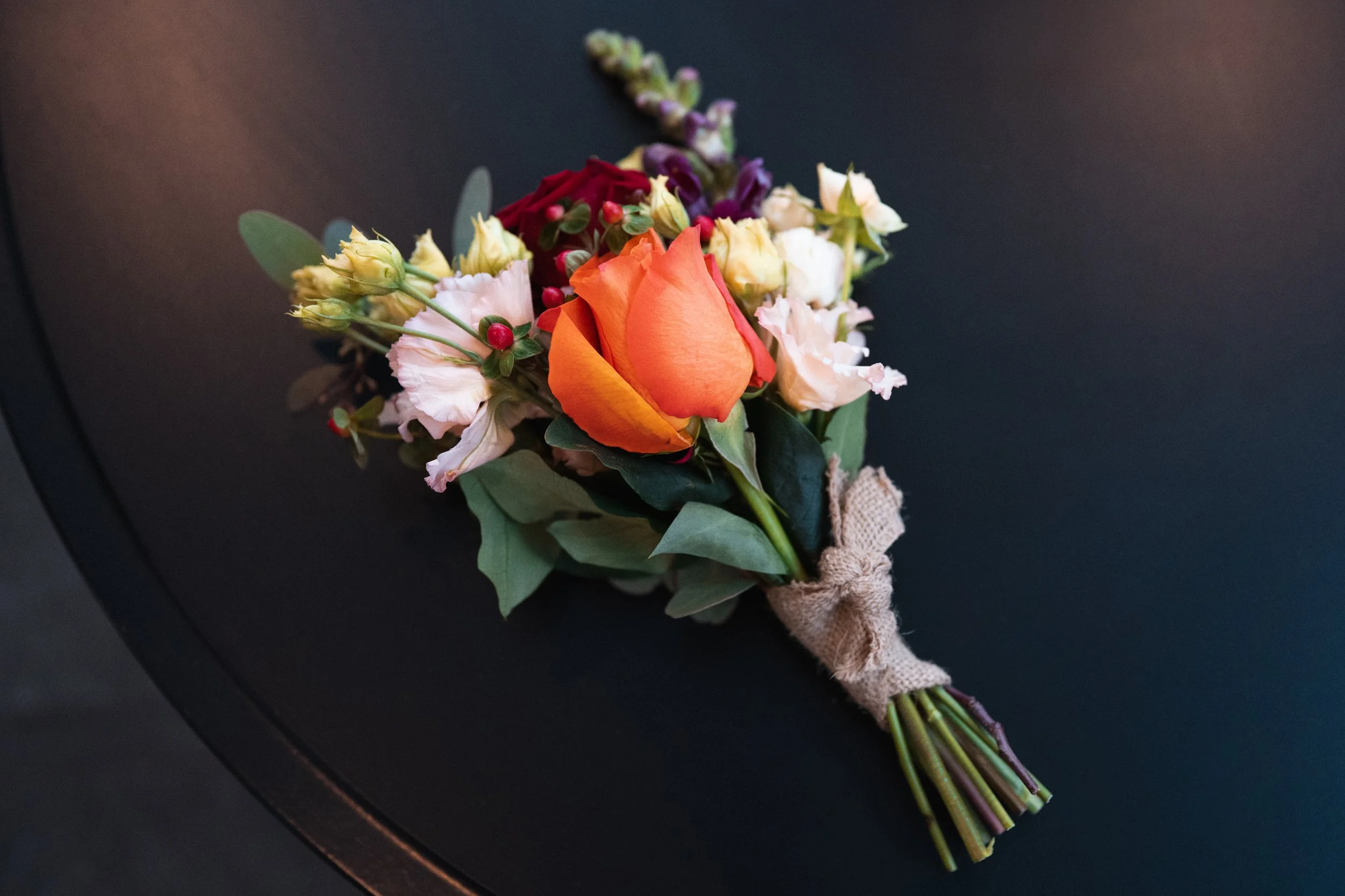A small bouquet of mixed flowers including a prominent orange rose, pink and white flowers, green leaves, and red berries, wrapped with burlap.