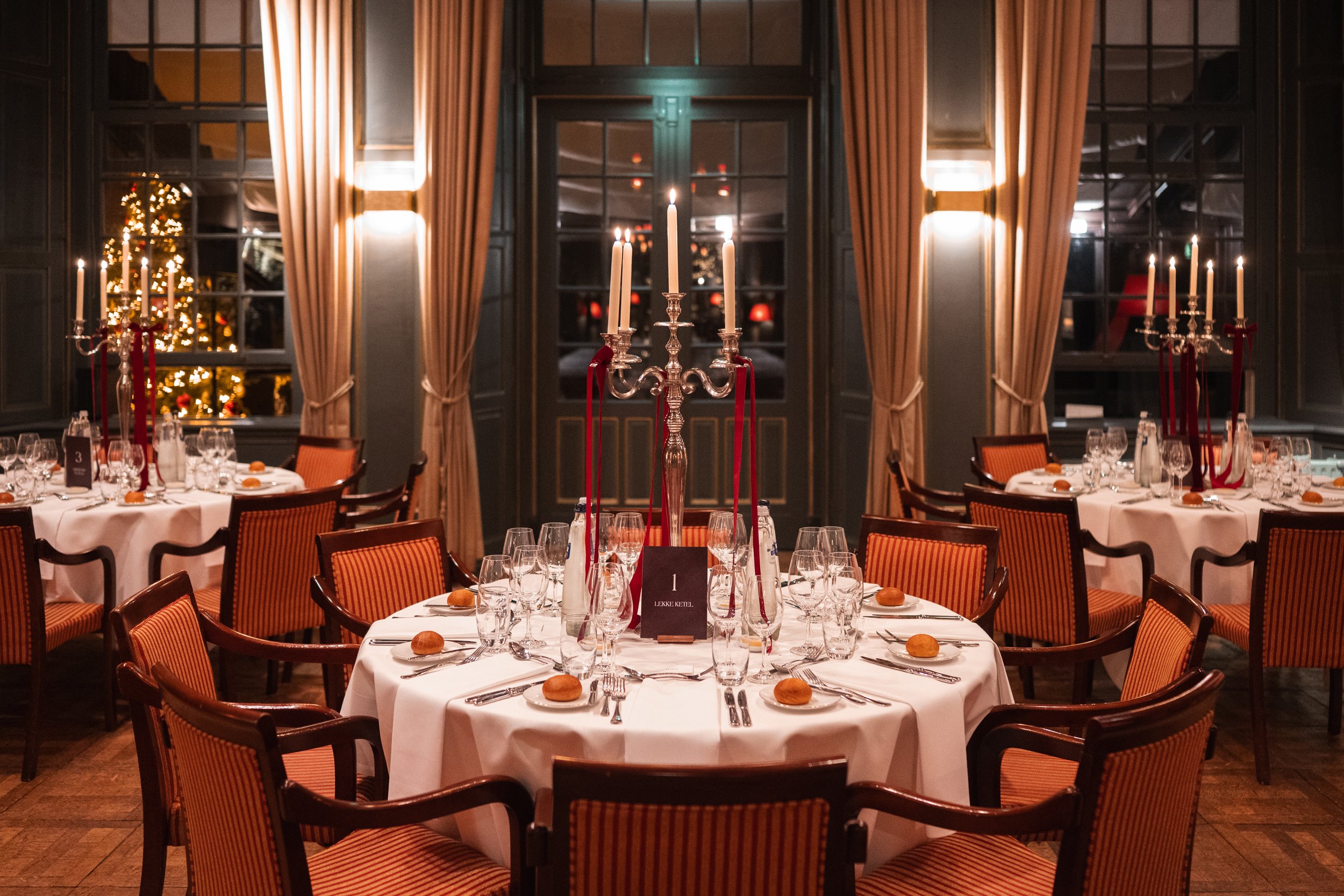 Elegant dining room with round tables covered in white tablecloths, set with glasses, silverware, and small bread plates, decorated with tall silver candelabras with white candles and red ribbons, in front of large windows framed by beige curtains, w