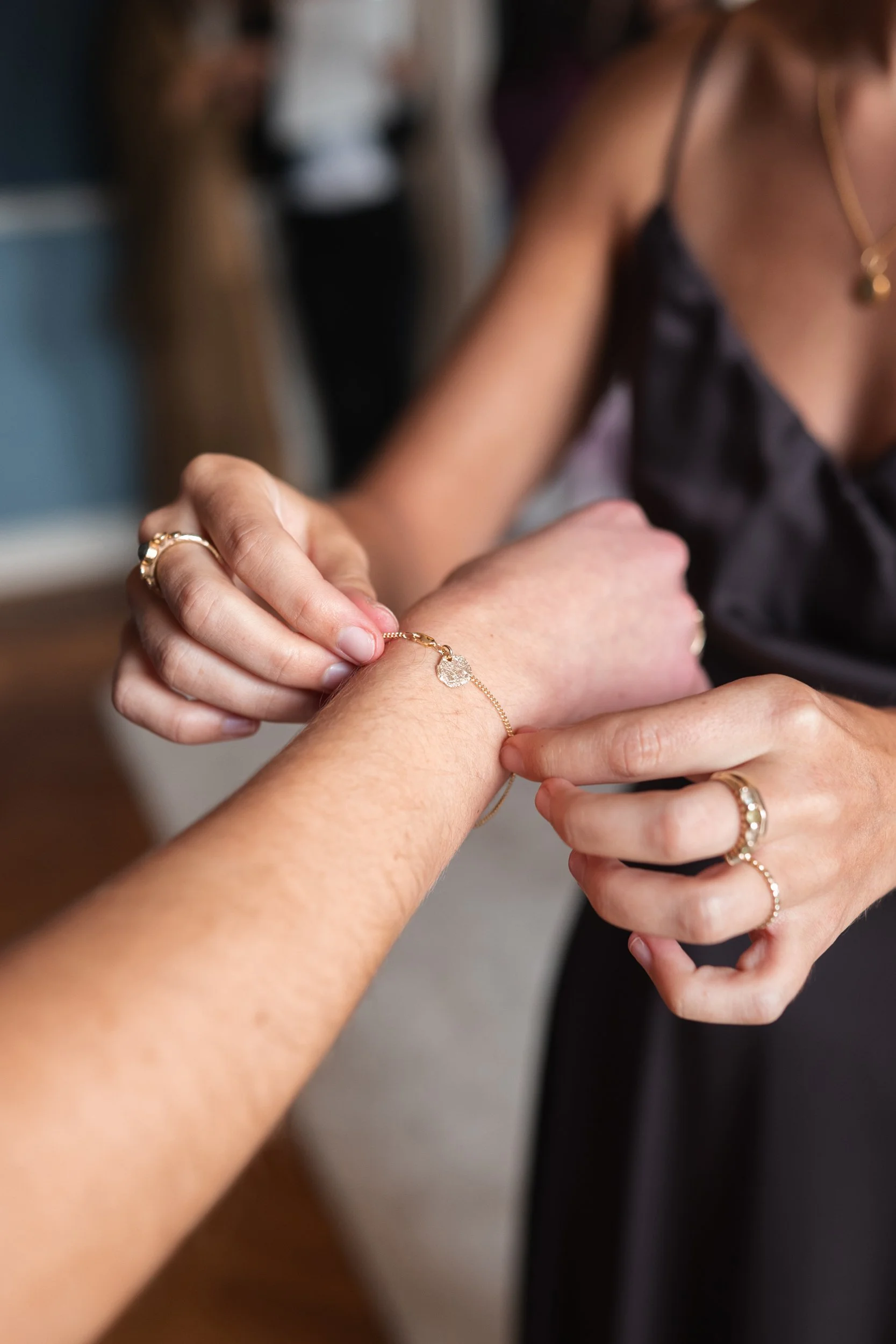 A woman holding and placing a bracelet on another person's wrist, both wearing gold rings, jewelry, and black clothing, with a blurred background.