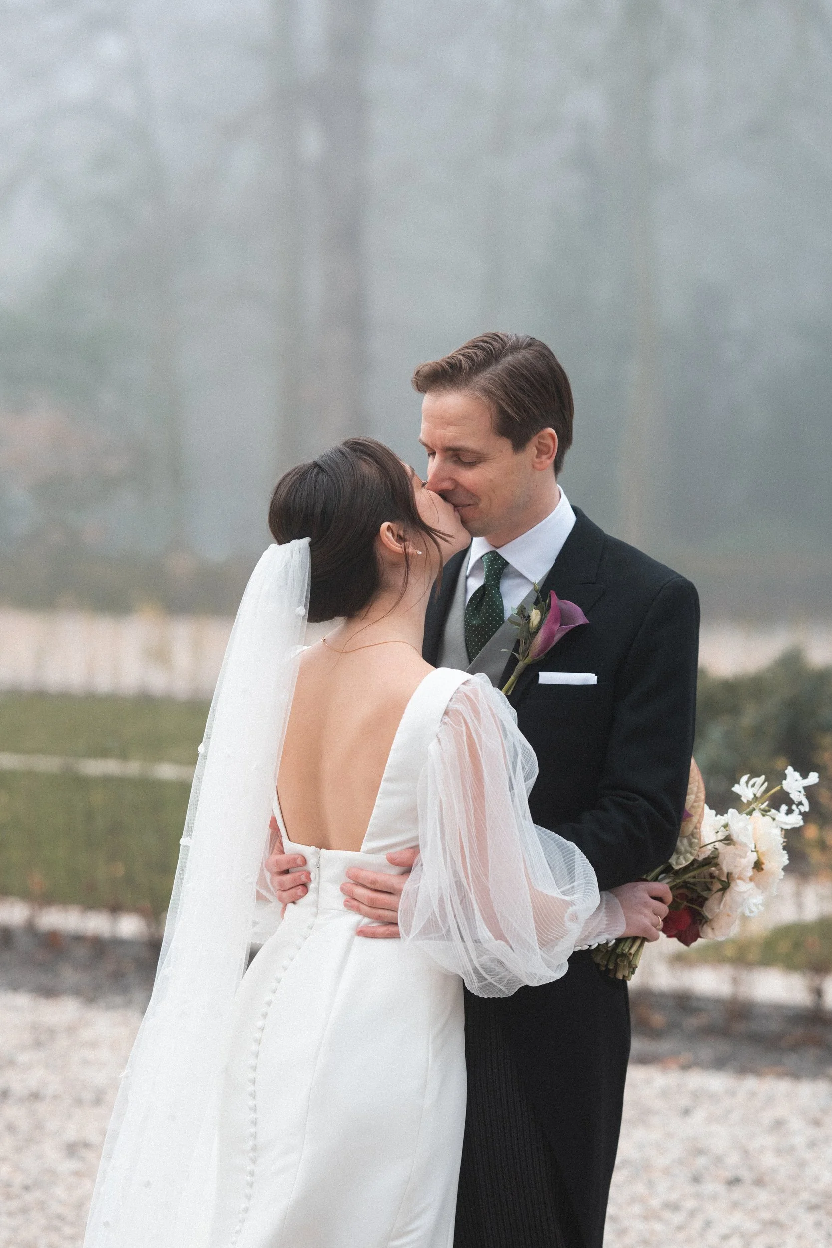 A couple in wedding attire sharing a kiss outdoors on a foggy day, with the woman in a white wedding dress and the man in a black suit holding a bouquet of flowers.
