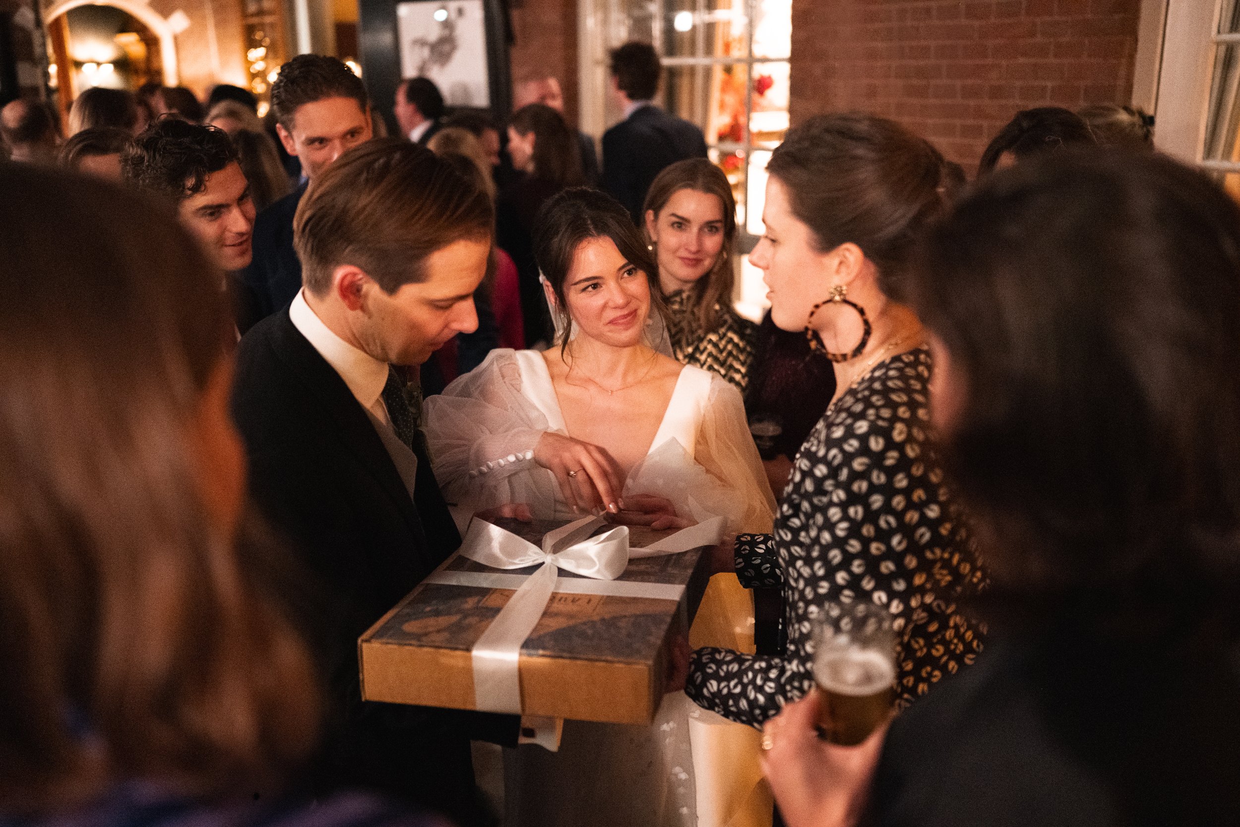 A woman in a white dress is giving a gift to a man in a suit at a social gathering, surrounded by people in conversation and holding drinks, in a warmly lit indoor setting.