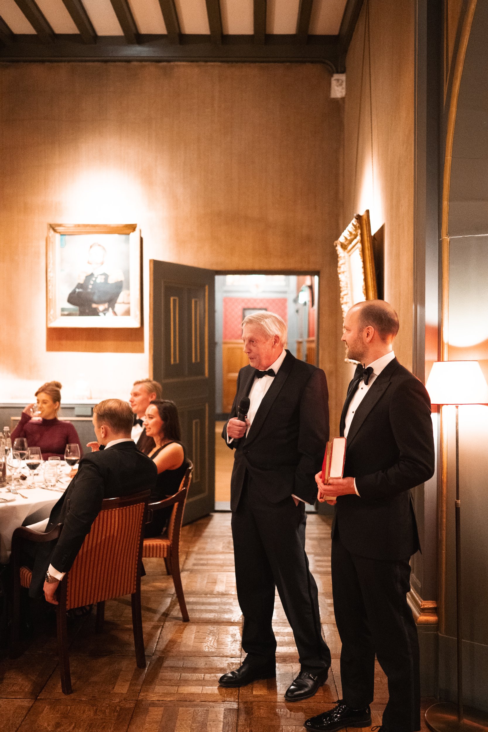 An older man in a tuxedo giving a speech, standing beside a younger man in a tuxedo holding a small gift, at a formal dinner event with guests seated at a table.