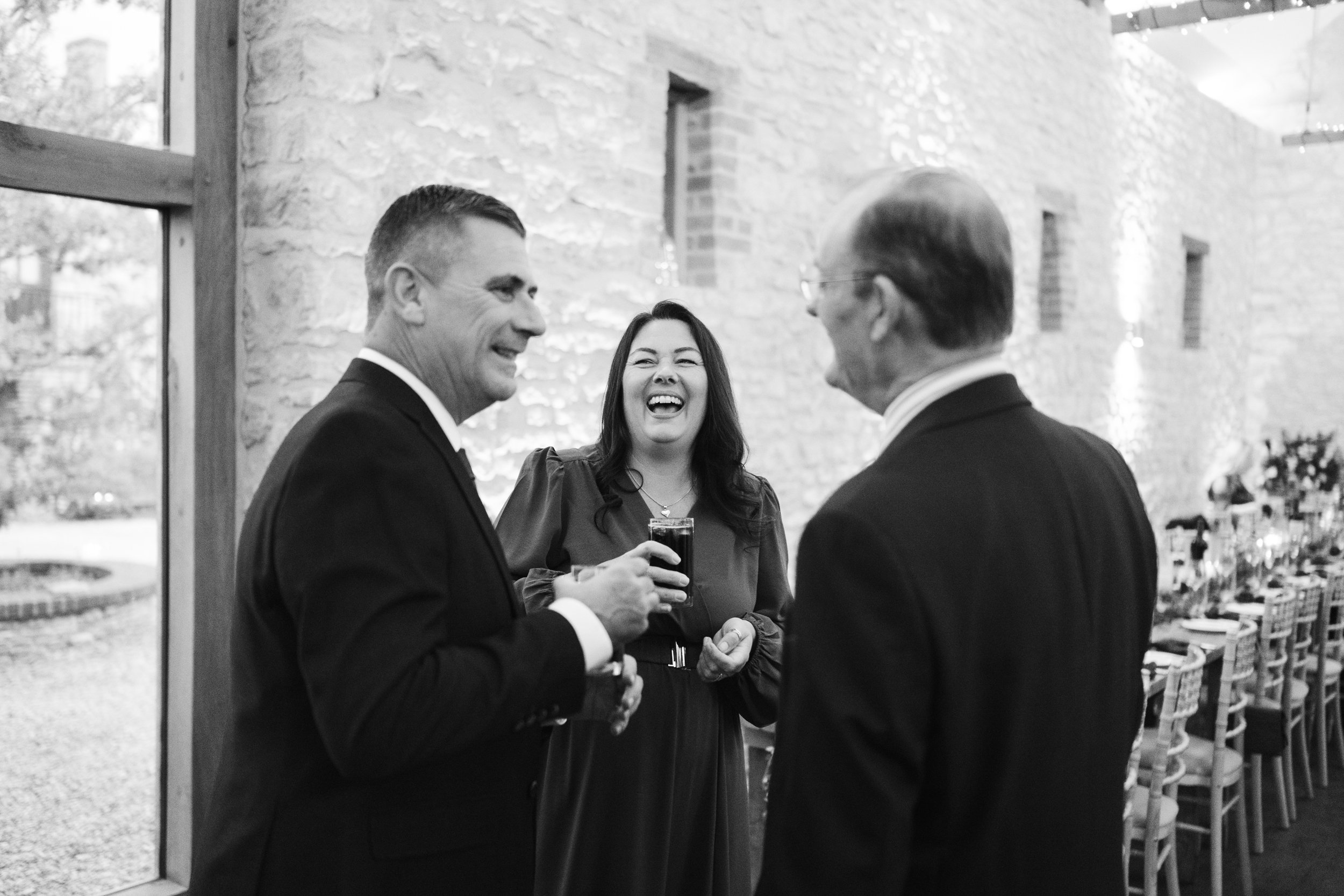 Three people in formal attire engaged in conversation at an indoor event, with one woman laughing.
