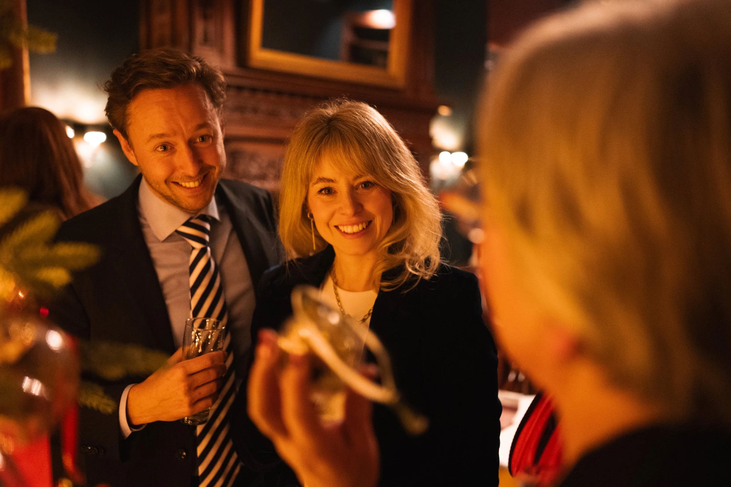 Two smiling people, a man and a woman, enjoying a social gathering, holding drinks, with blurred figures and warm lighting in the background.