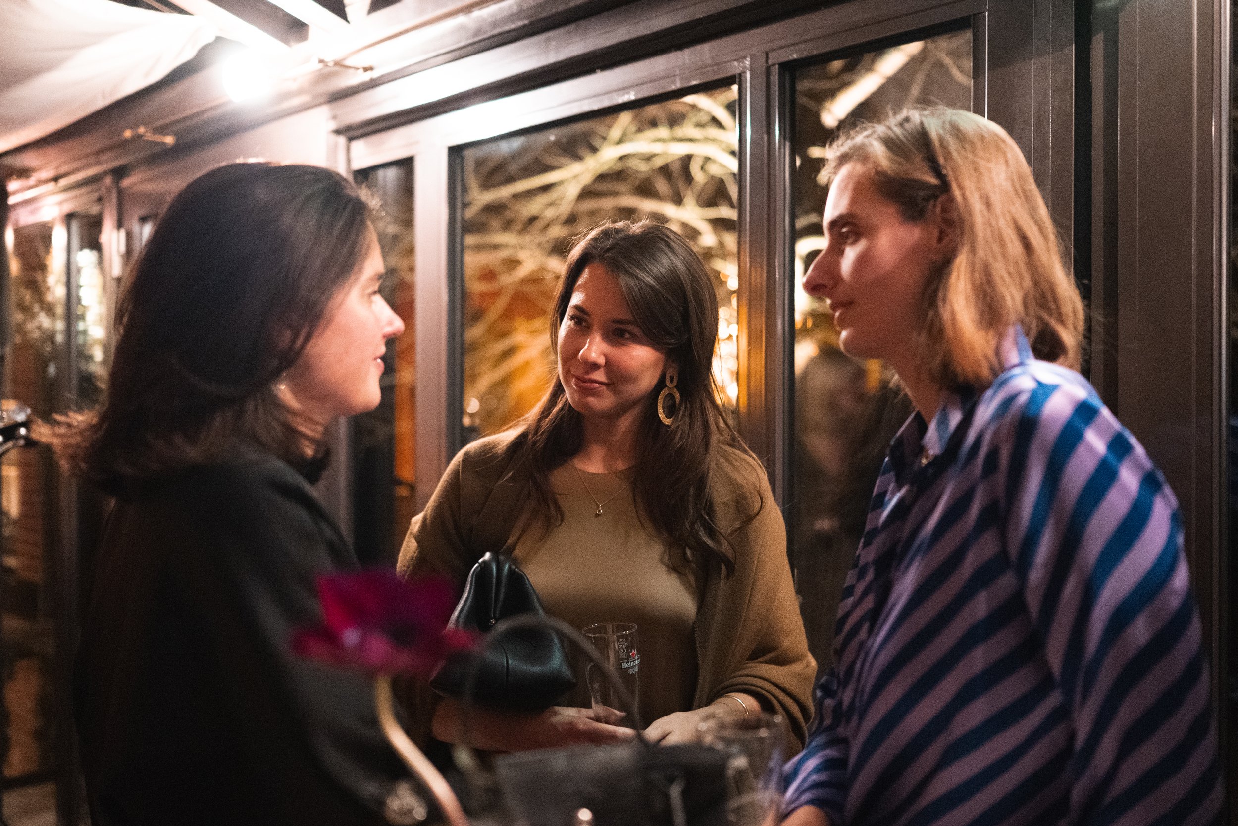 Three women are engaged in conversation at a social gathering indoors at night, with warm lighting and large windows behind them.