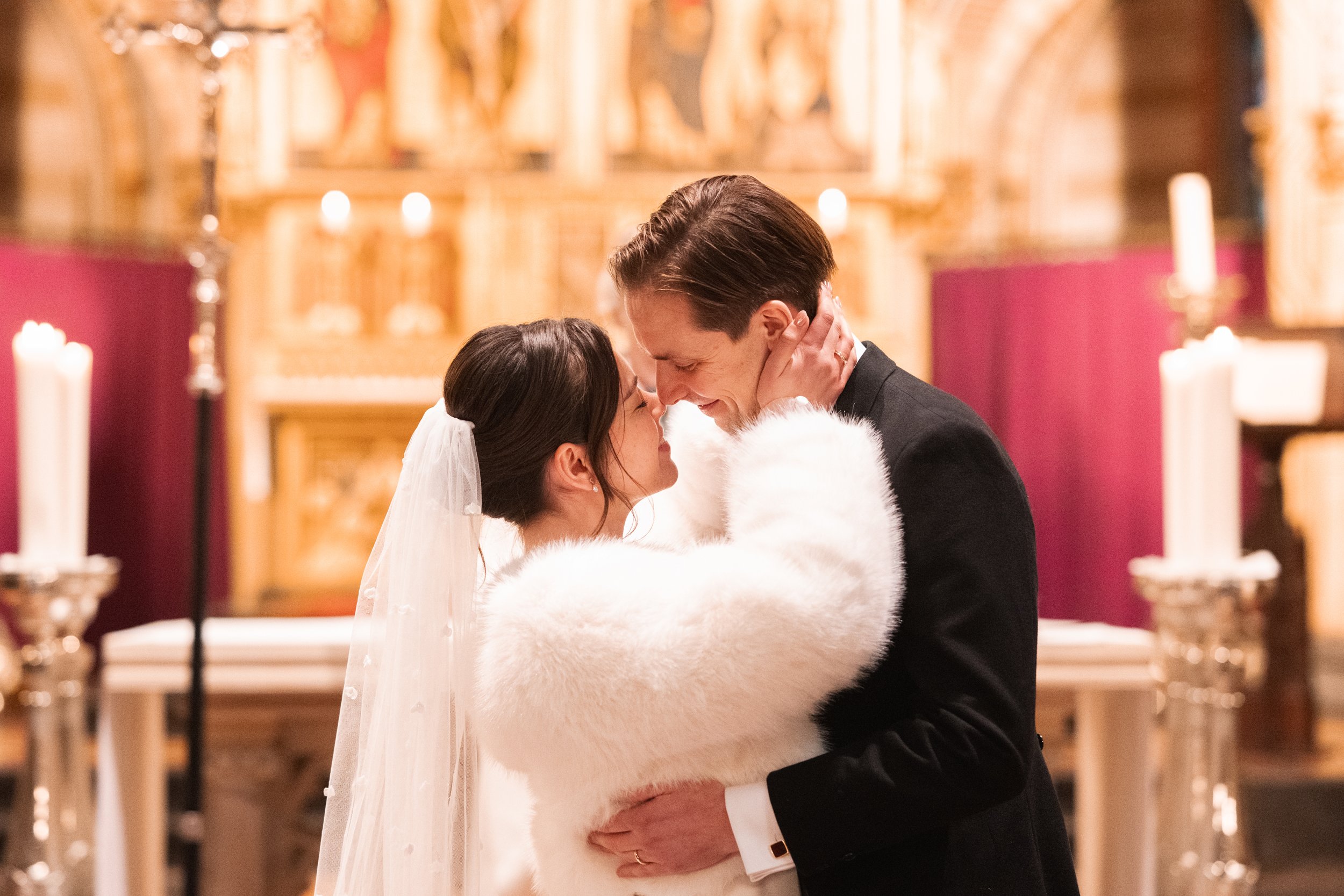 A bride and groom share an intimate moment during their wedding ceremony inside a church, with candles and ornate decorations in the background.